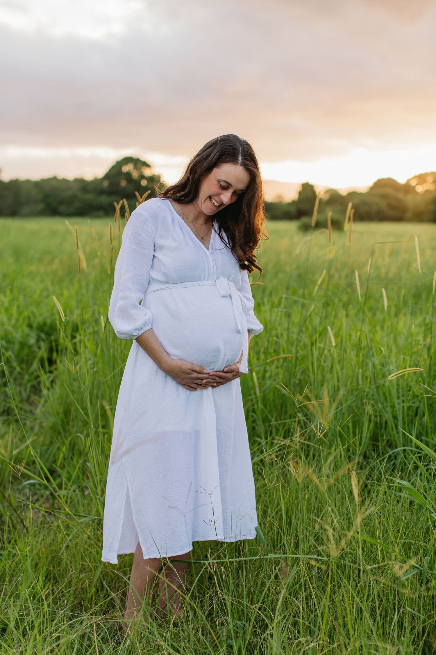 Pregnant woman in white dress cradling baby bump in green field at sunset.