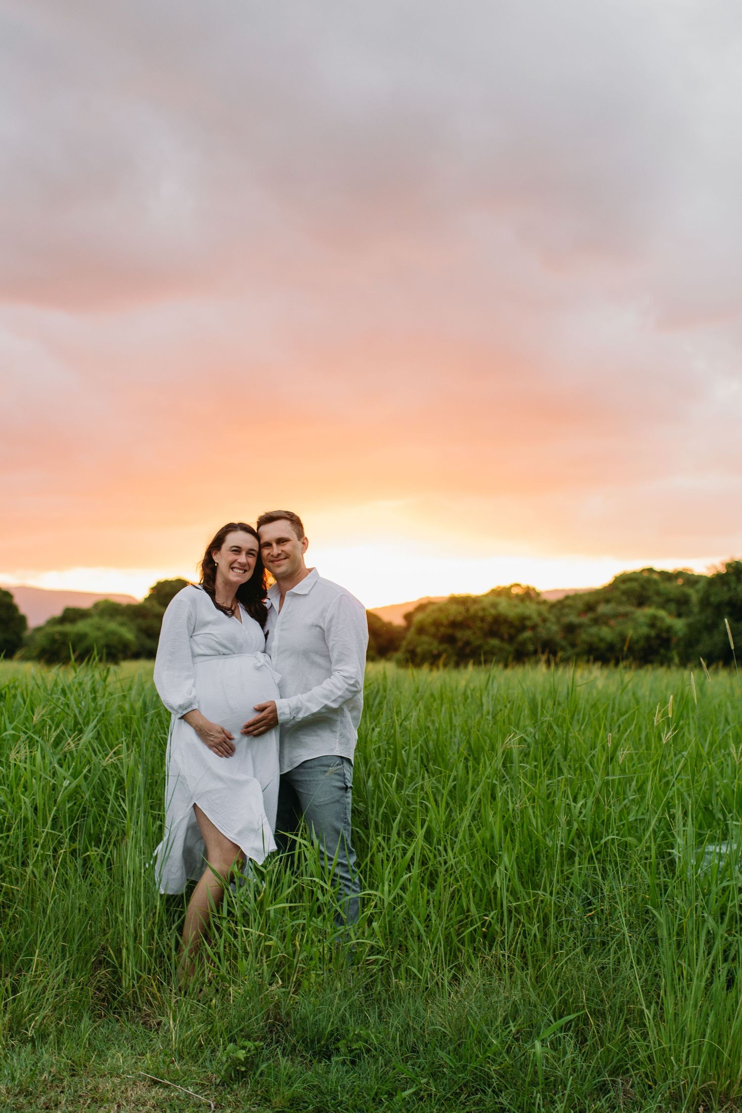 Couple poses in green field at sunset, woman in white dress cradling baby bump, man in white shirt embracing her.