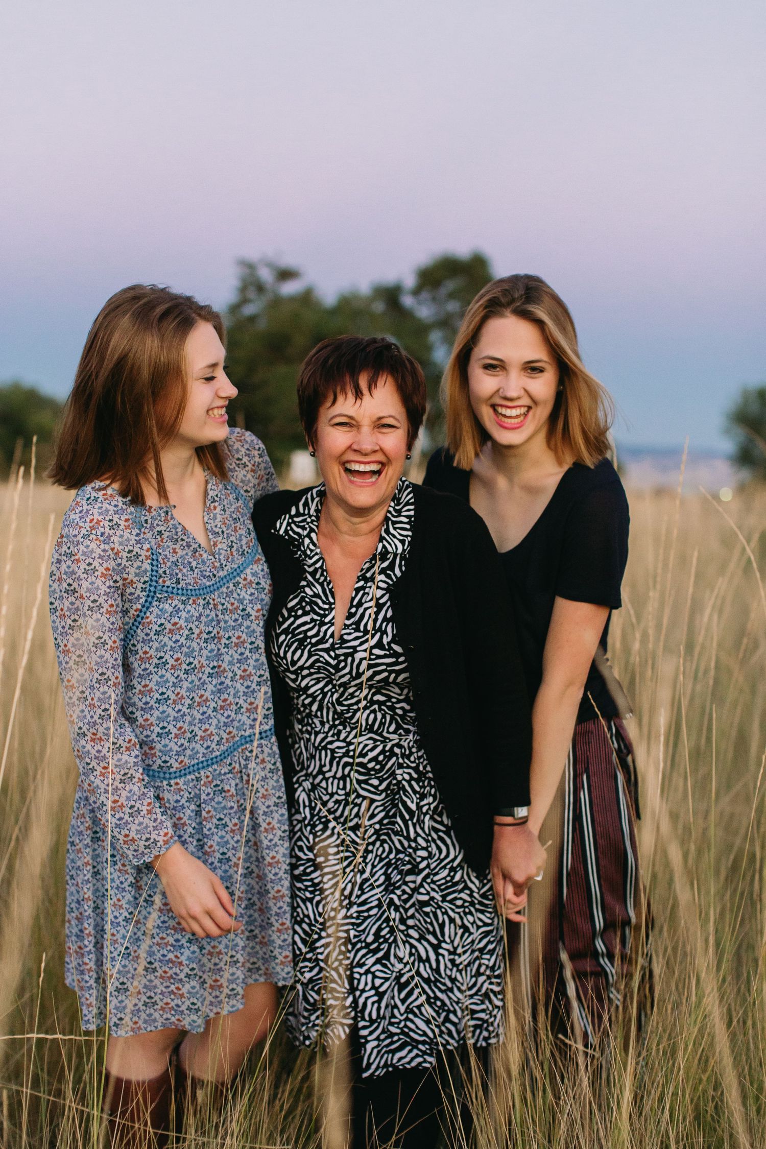 Three smiling women standing together in a golden grass field at dusk, wearing casual dresses.