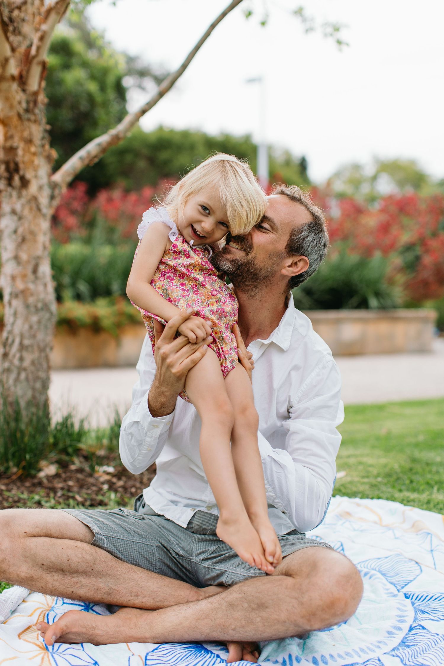 Father sitting cross-legged on blanket in park, kissing young blonde daughter in floral dress.