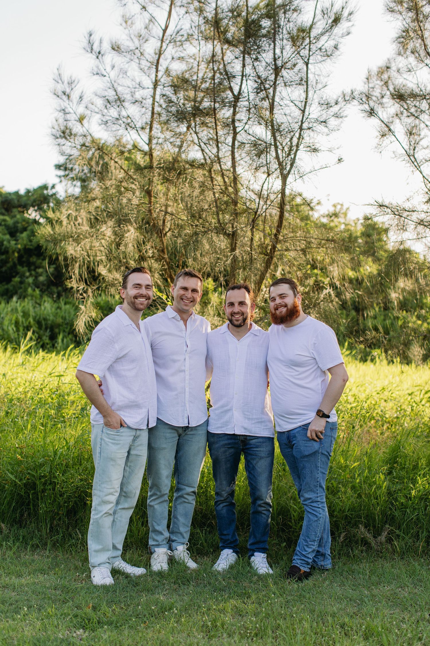 Four smiling men in white shirts and jeans pose together outdoors in a lush green park setting.