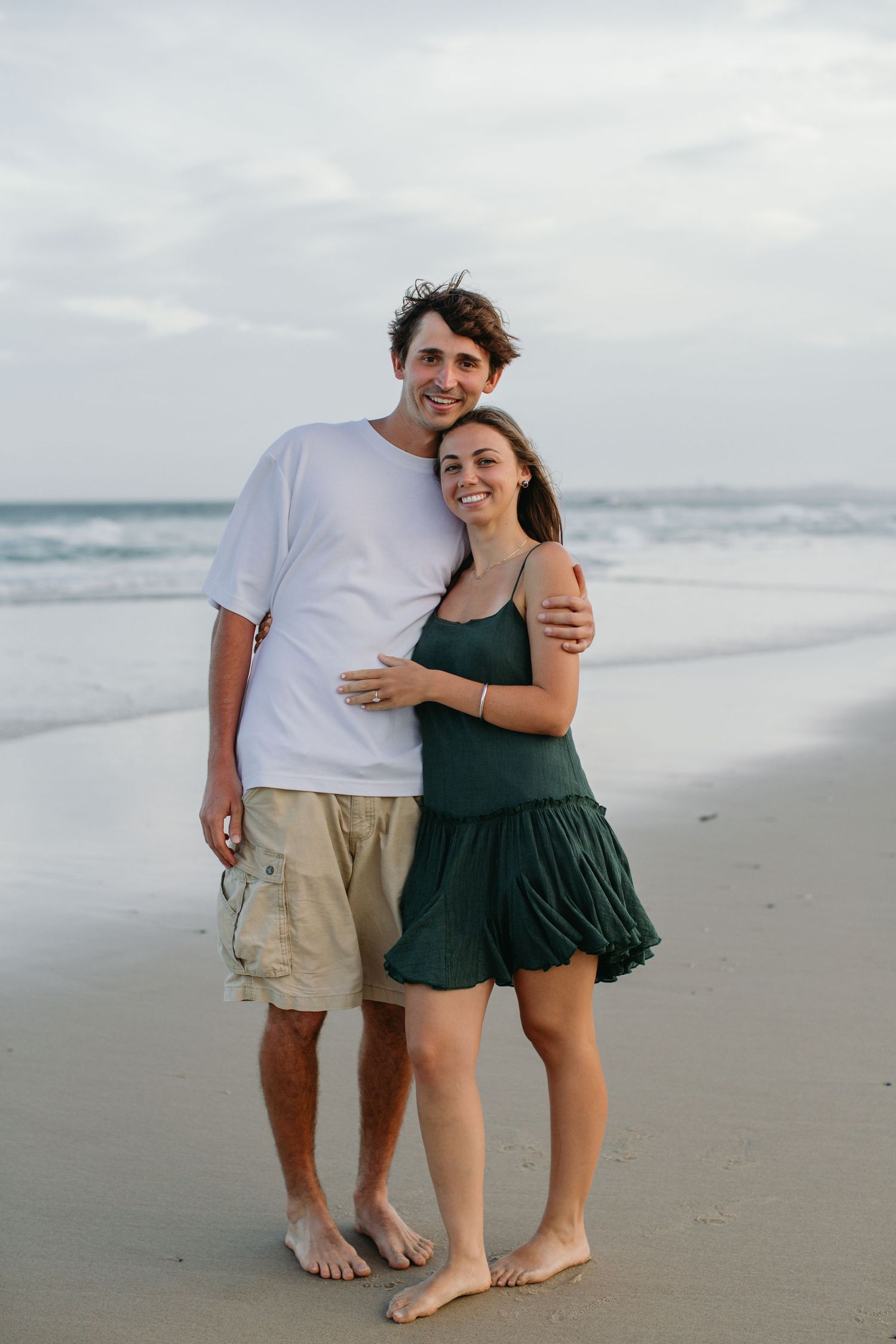 Happy couple embracing on a sandy beach, man in white tee and khaki shorts, woman in green dress, ocean waves behind.