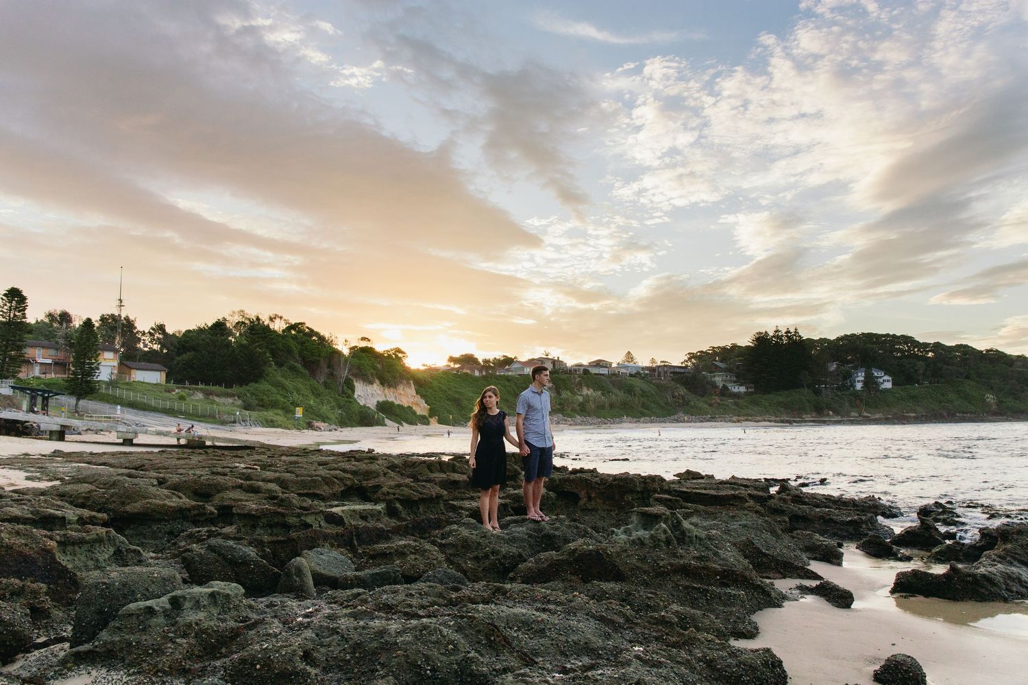 Couple standing on rocky shoreline at sunset with ocean waves and lush green headland in background.