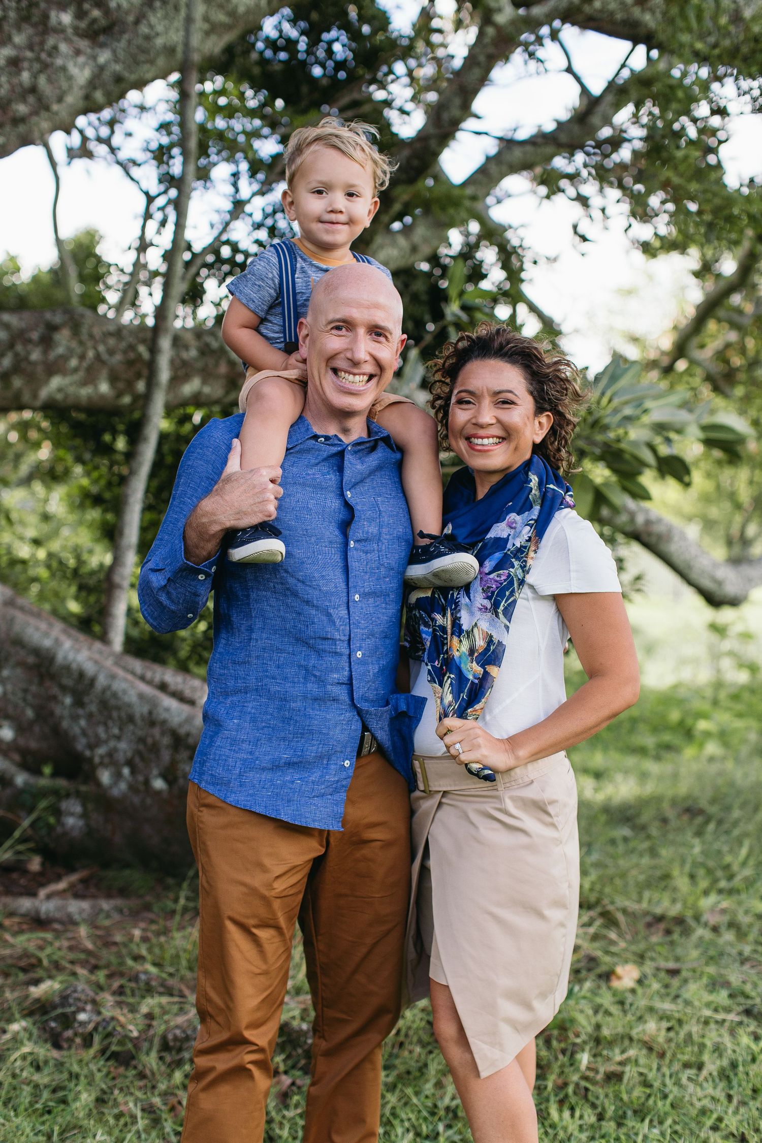 Happy family of three posing outdoors, father carrying child on shoulders with mother beside him in lush green park.