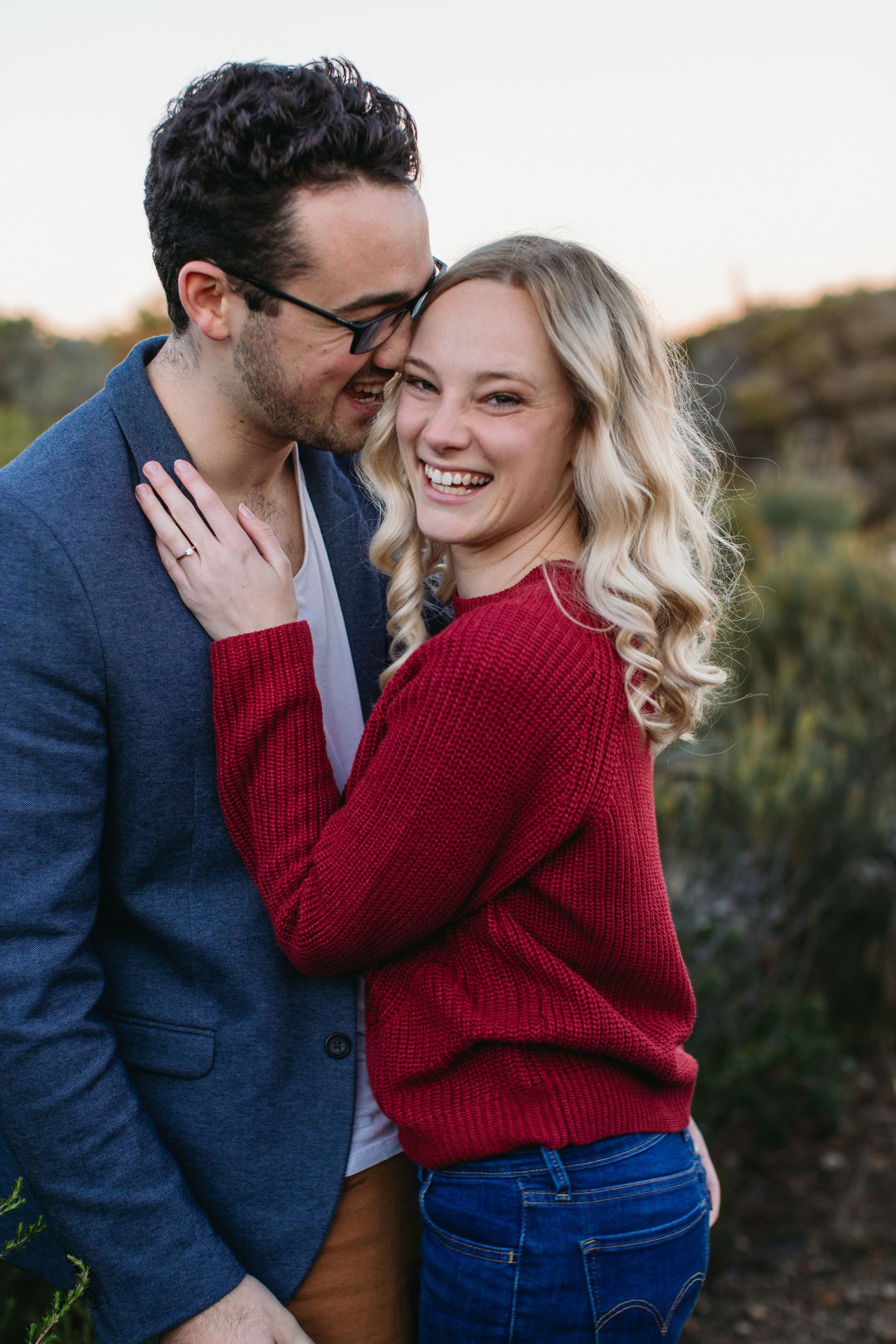 Happy blonde woman in red sweater laughing as man in blue blazer embraces her.