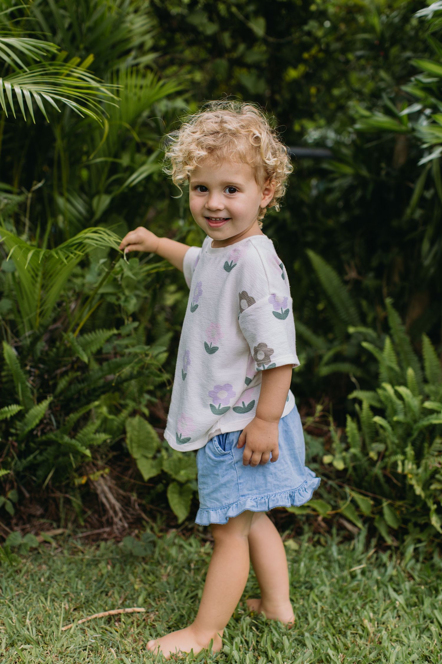 Curly-haired toddler girl in floral top and denim skirt smiling in a lush green garden.