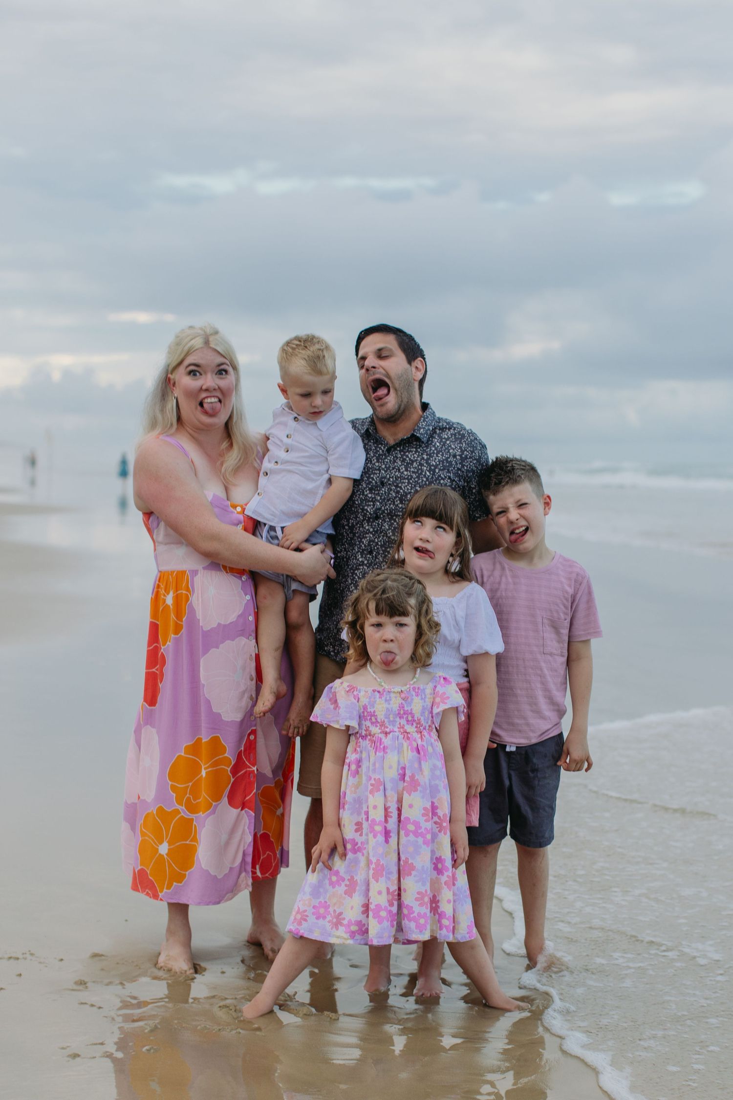 Family of six posing playfully on a beach, parents with four young children making funny faces by the ocean.