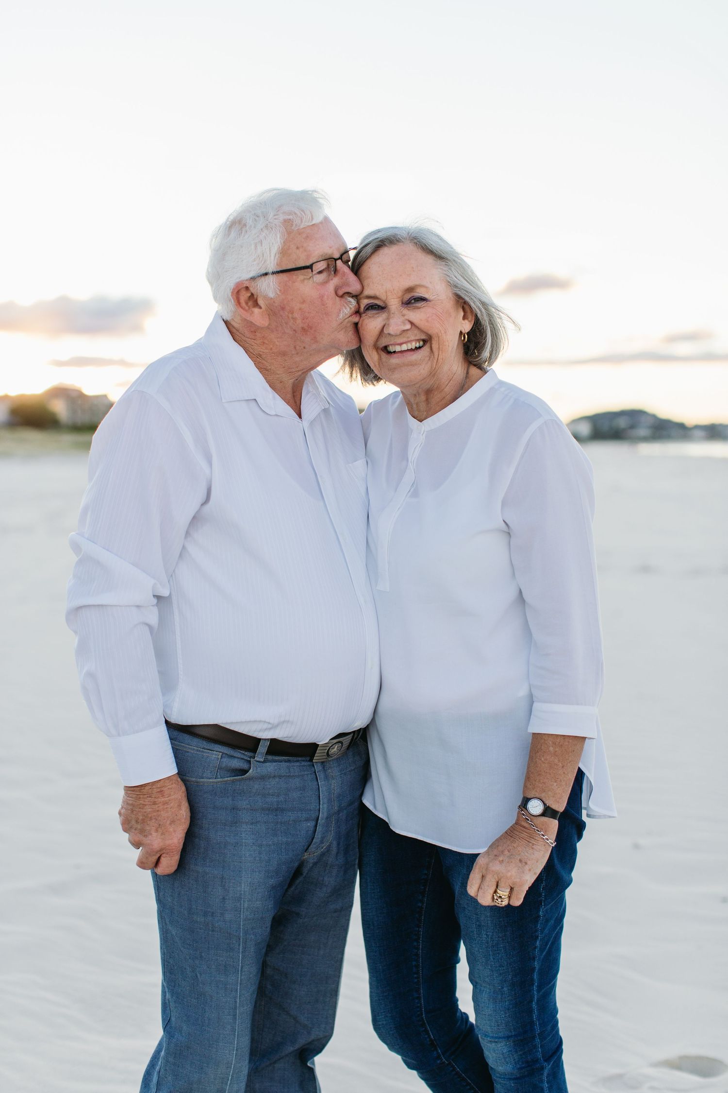 Elderly couple in white shirts and jeans sharing a kiss on the beach at sunset.