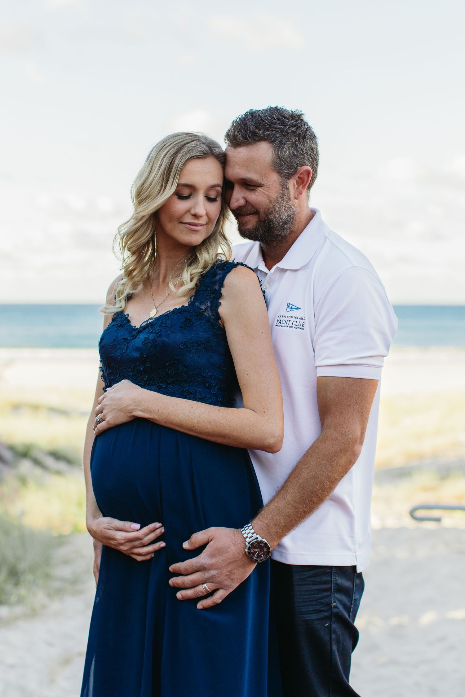 Couple shares tender maternity photo on beach, woman in navy lace gown cradling baby bump, man in white polo embracing her.