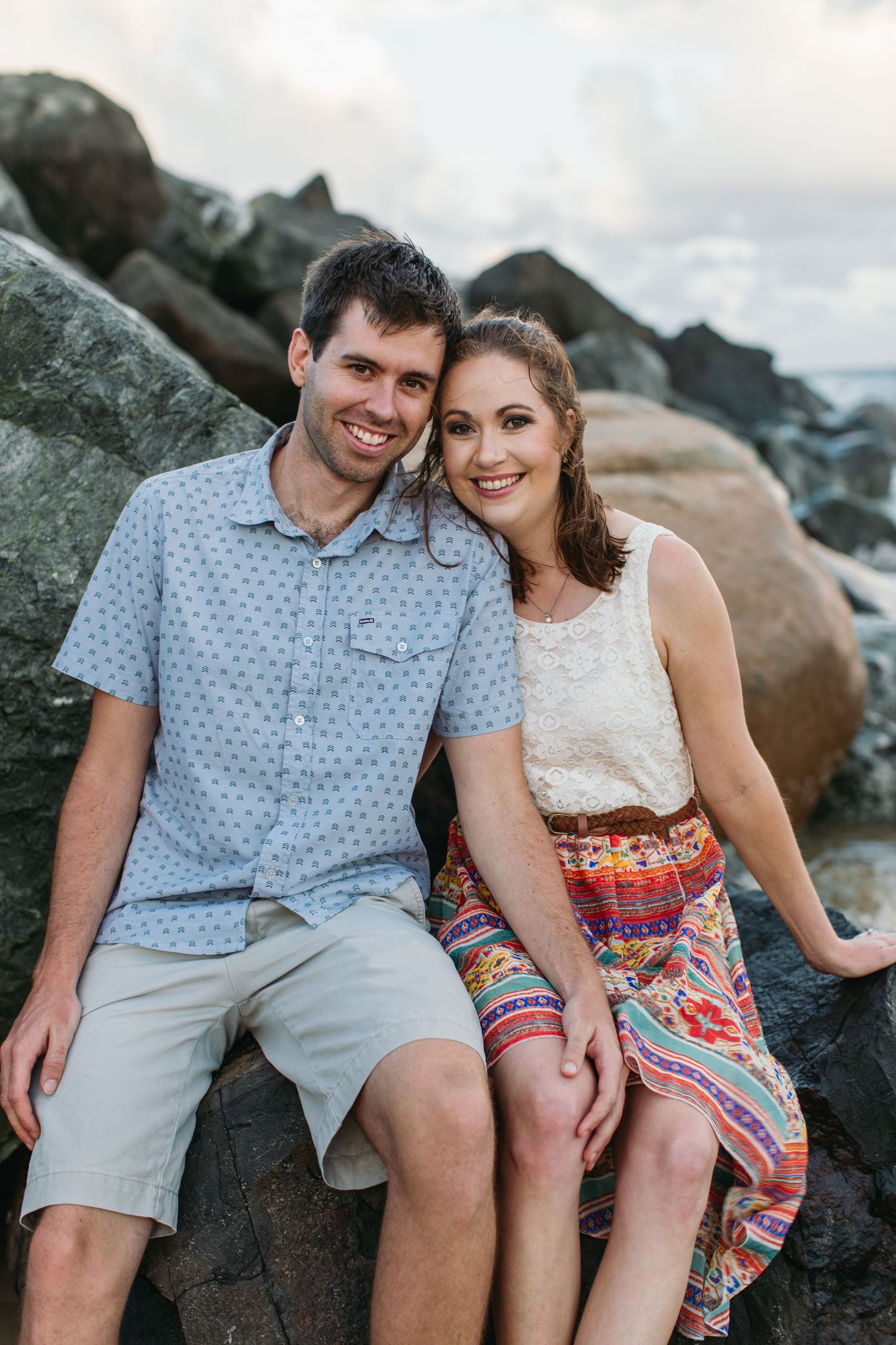 A smiling couple sits on rocky shore, woman in colorful skirt and man in blue shirt, posing for a portrait.