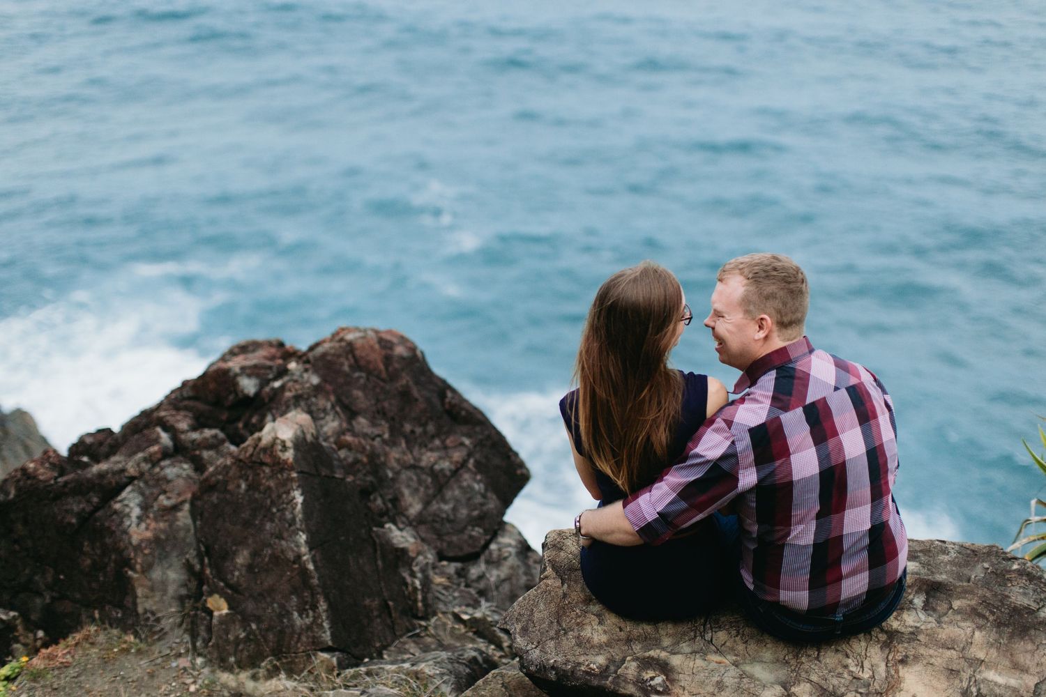 Couple sitting on rocky coastal cliffs embracing while gazing at turquoise ocean waves.