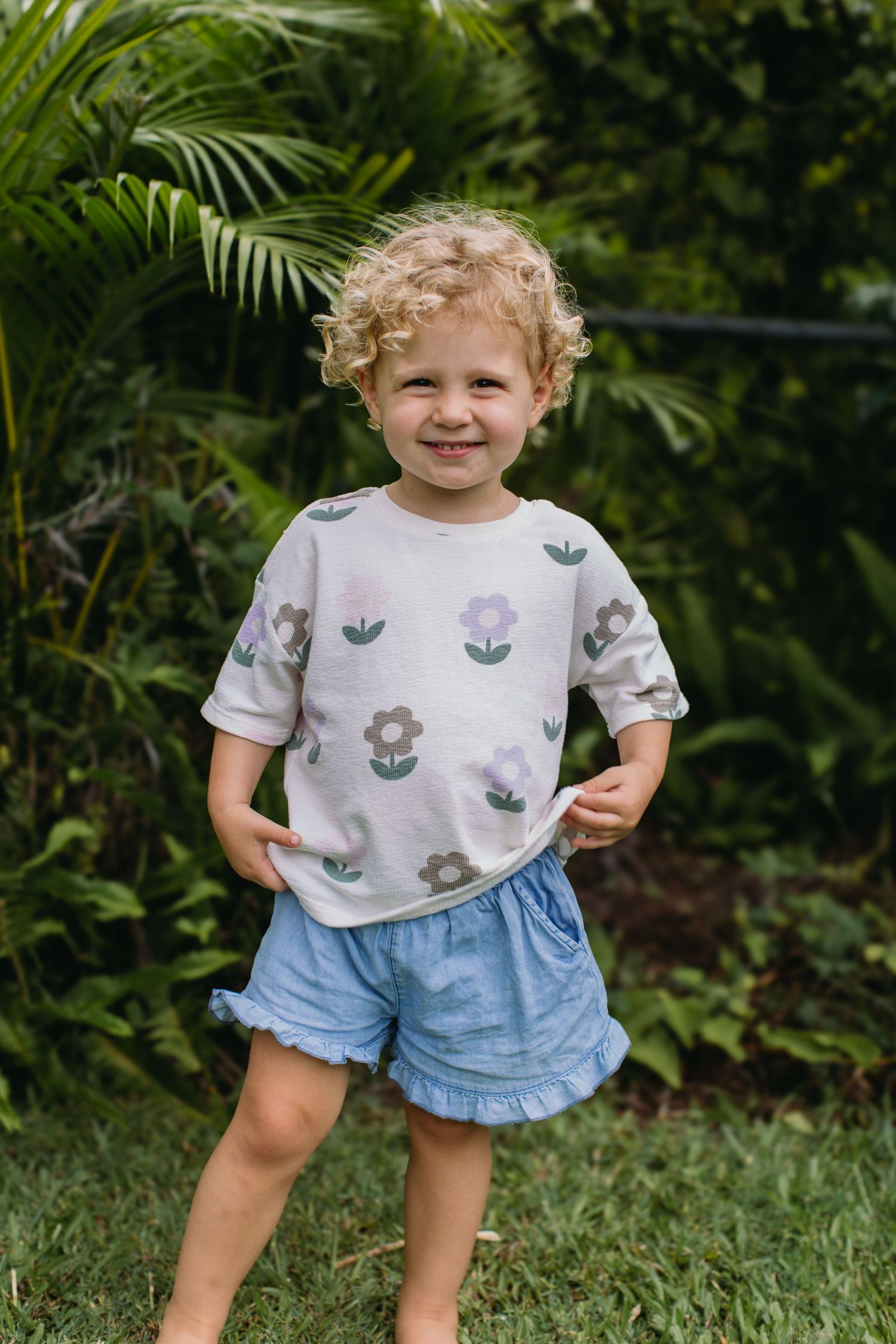 Smiling toddler with curly hair wearing a floral top and blue shorts, standing outdoors in a lush green garden.