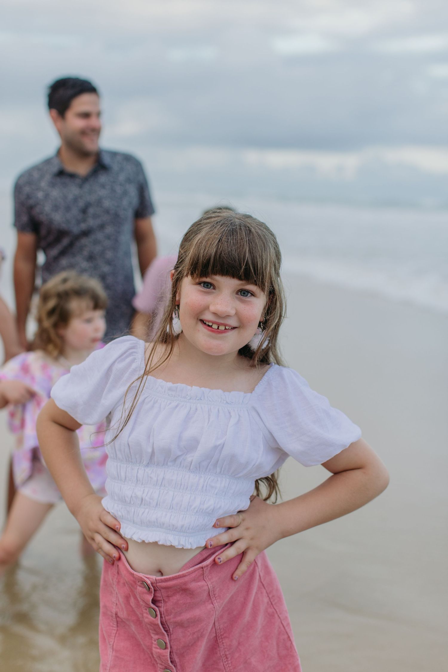 A cheerful young girl in a white crop top and pink skirt poses on the beach with family behind her.