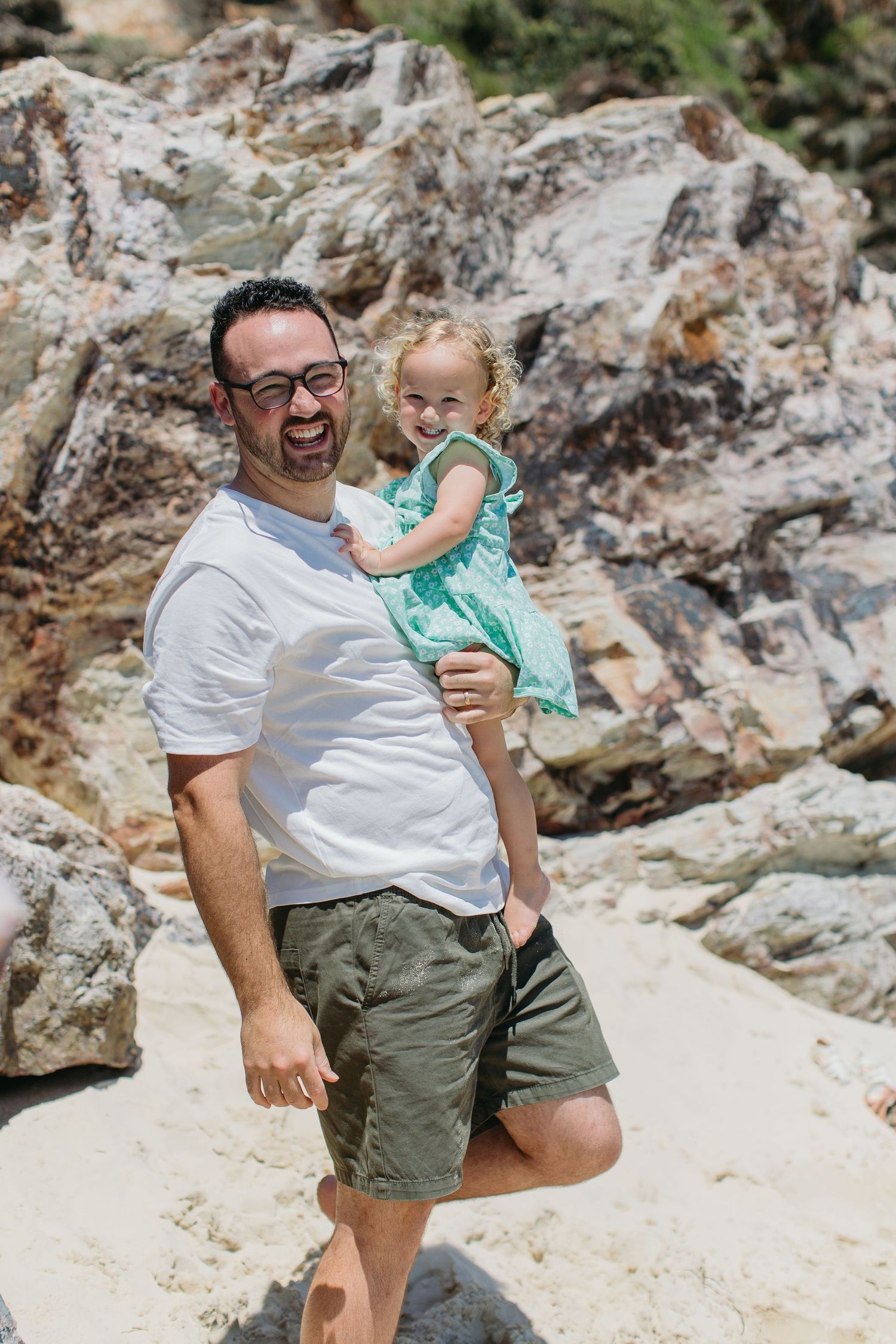 Laughing father carrying toddler girl in green dress at rocky beach on bright sunny day.