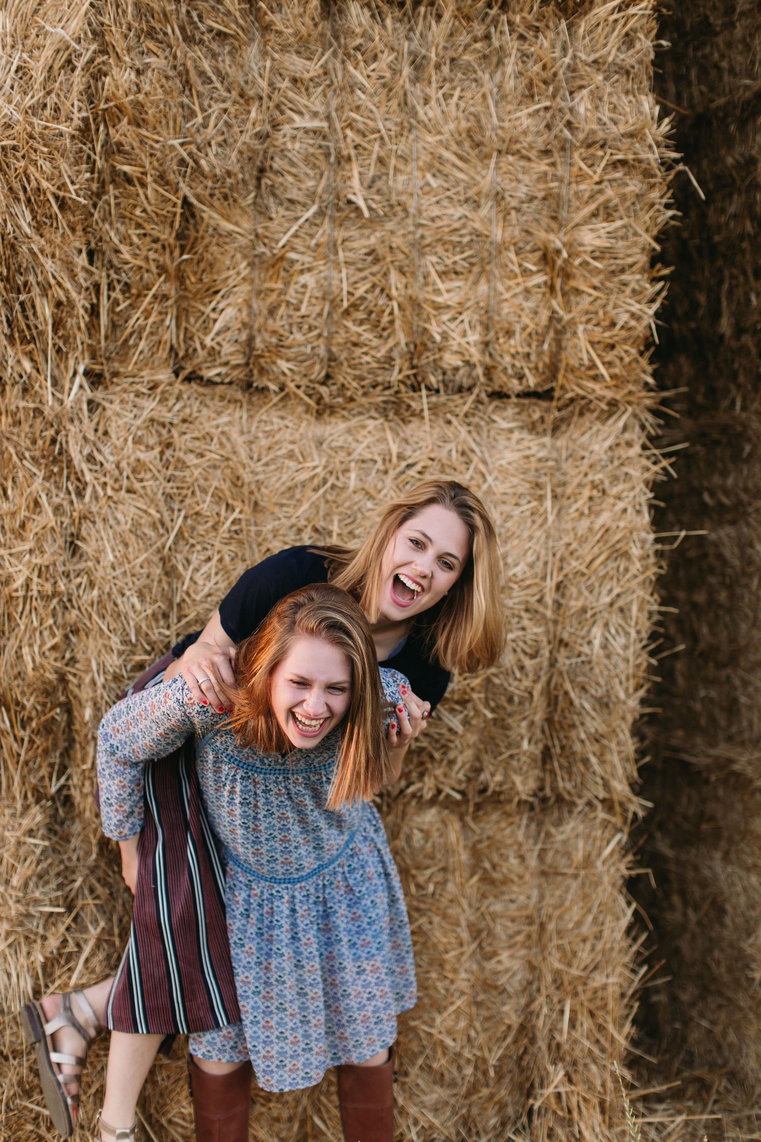 Two laughing young women pose playfully in front of stacked hay bales, one giving the other a piggyback ride.