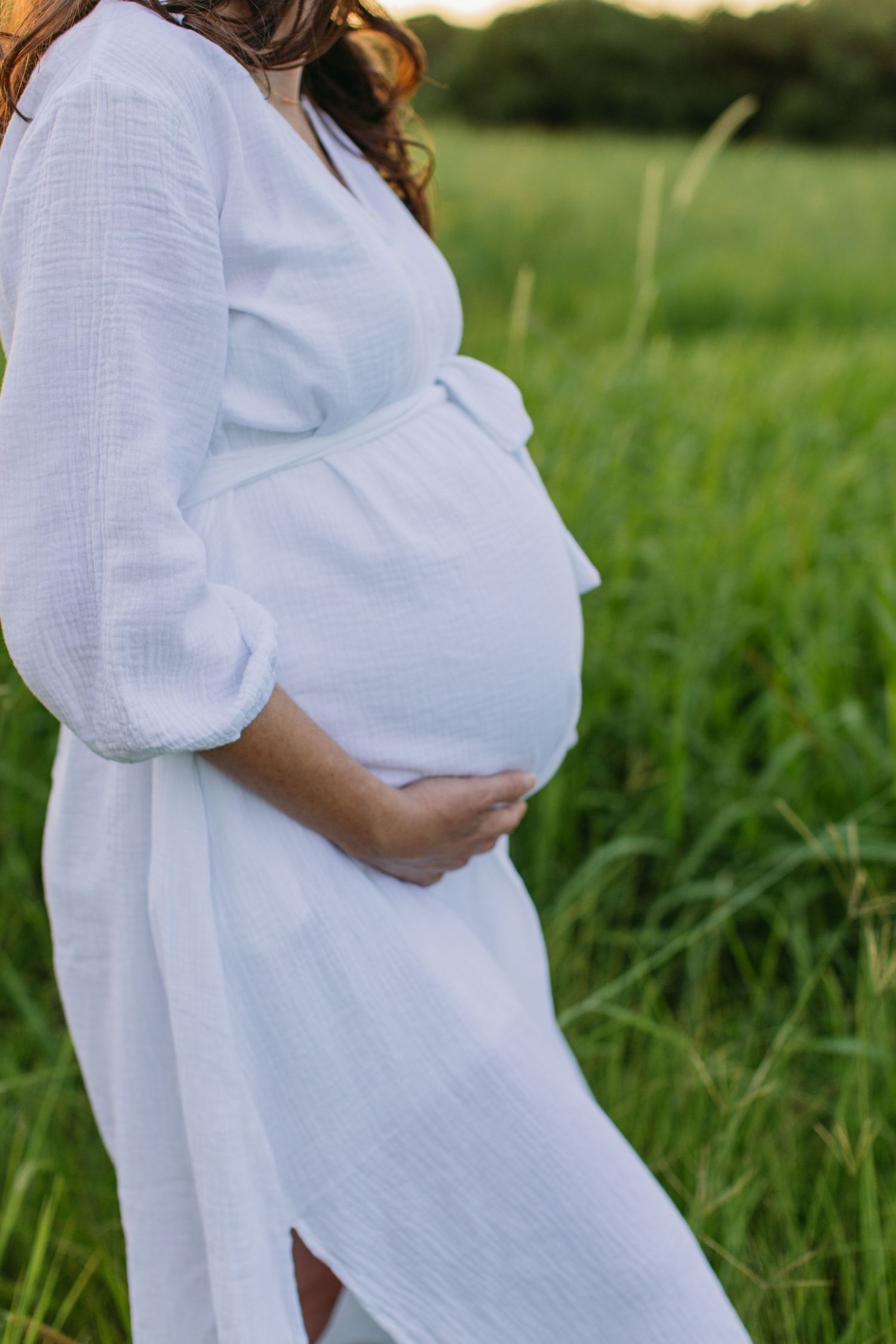 Pregnant woman in white linen dress holding baby bump in green field at sunset.