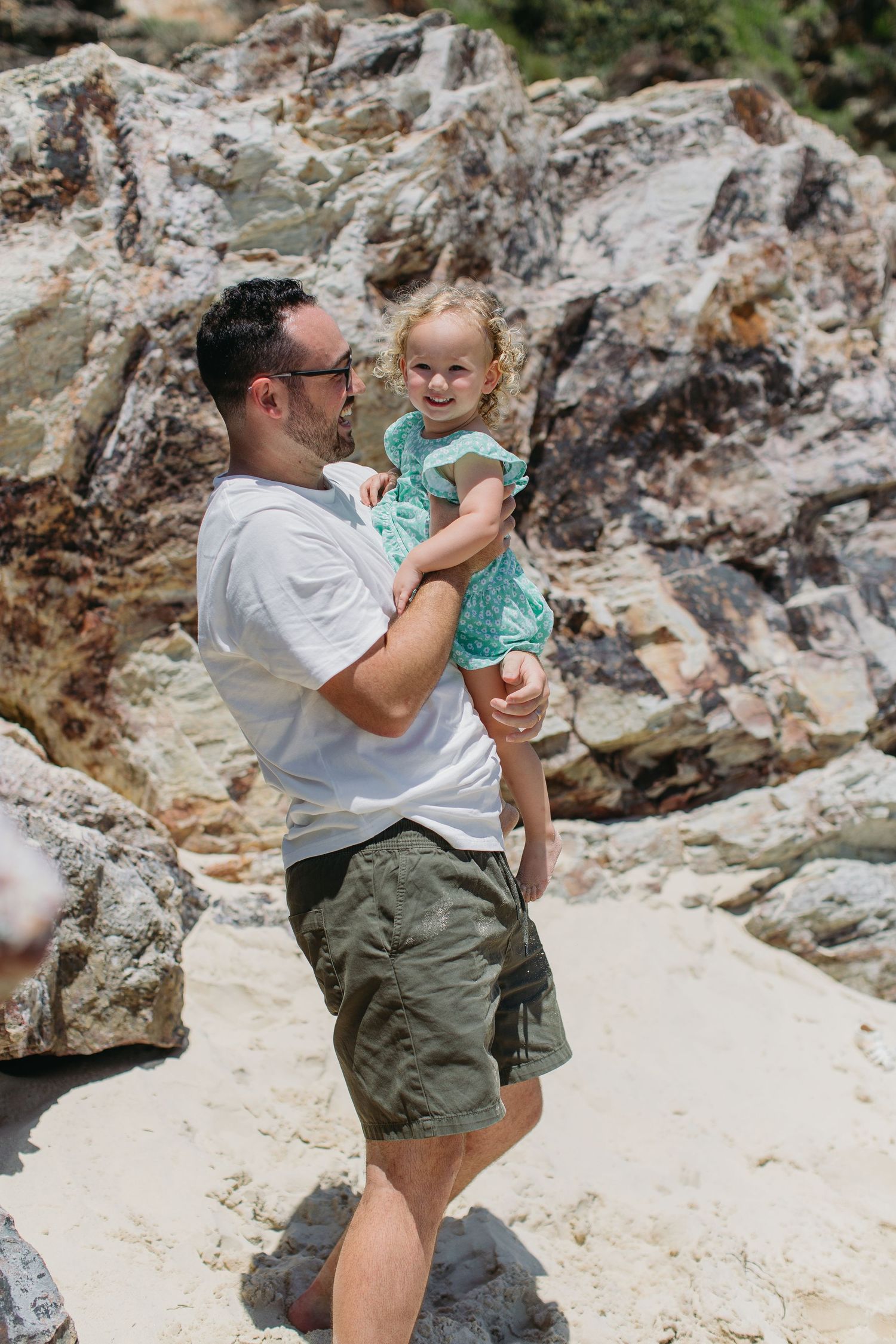 Man in white shirt and camo shorts holds smiling toddler girl in teal dress near rocky beach.