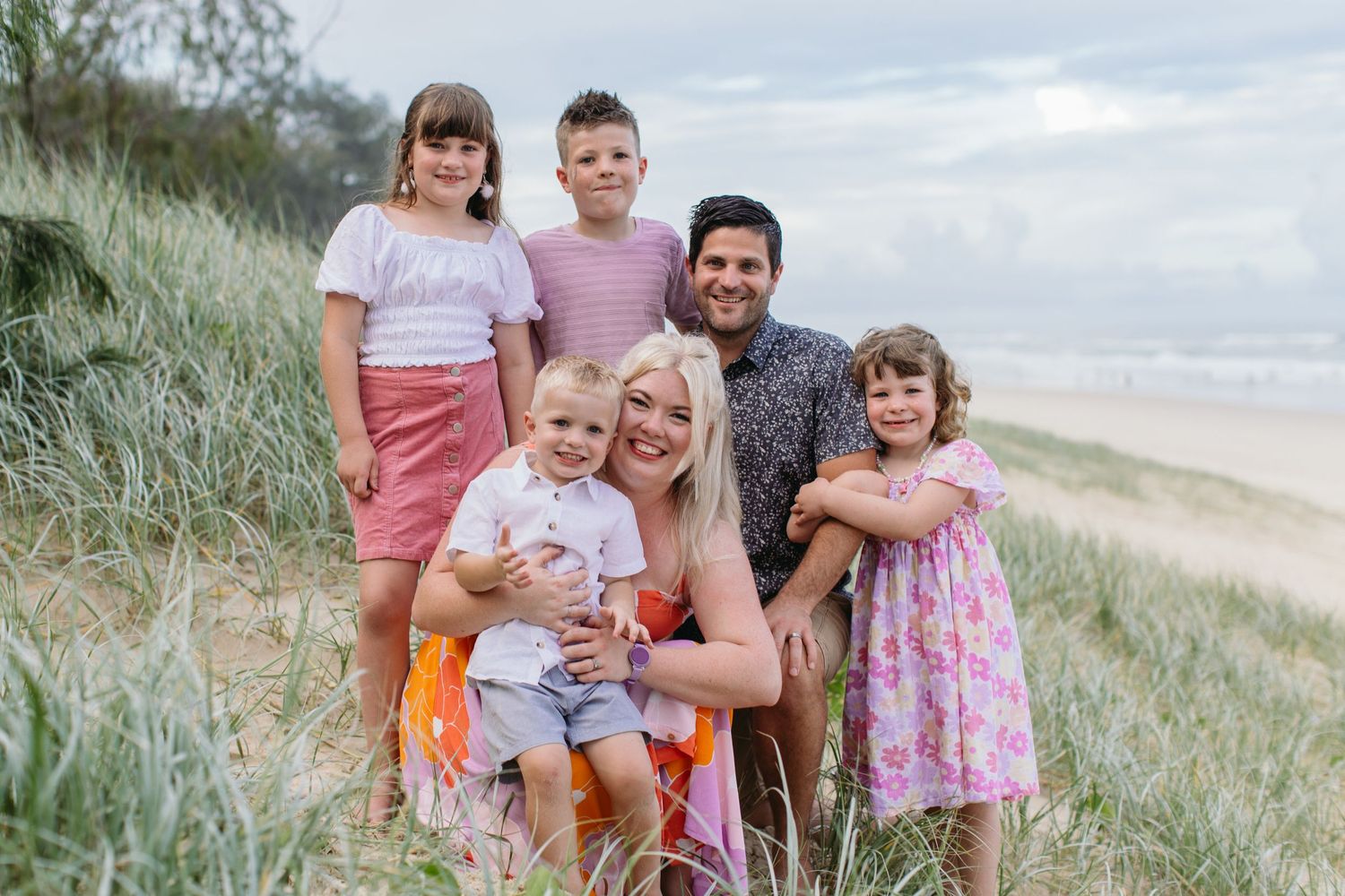 Family of six smiling together on a sandy beach among grass dunes on a cloudy day.