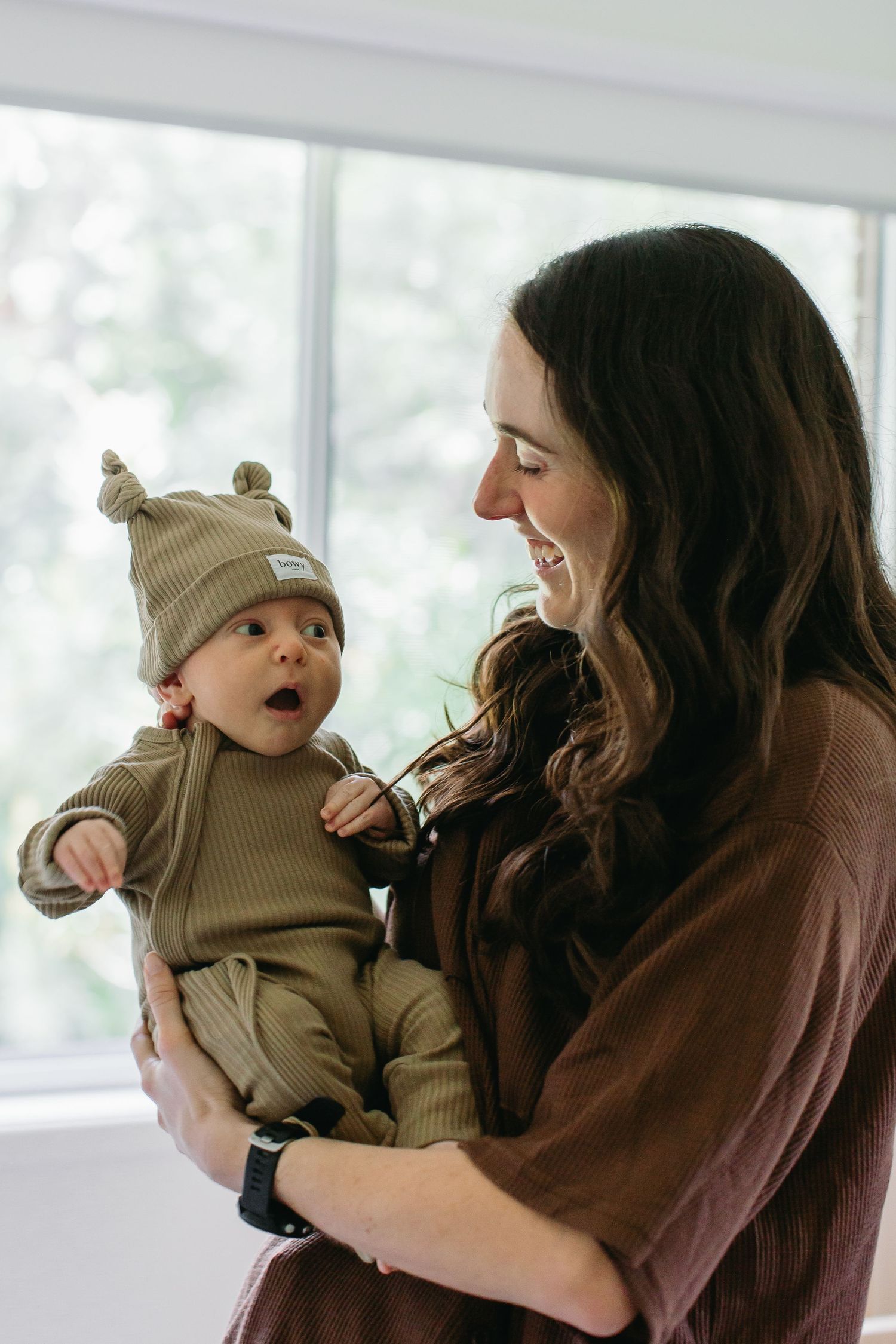 Smiling mother holding her wide-eyed baby dressed in a ribbed olive romper and knot beanie hat.