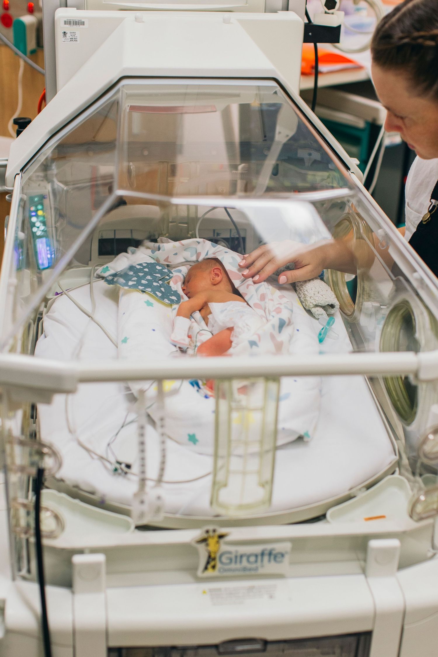 Newborn baby in a Giraffe incubator in the NICU with a nurse gently caring for the infant.