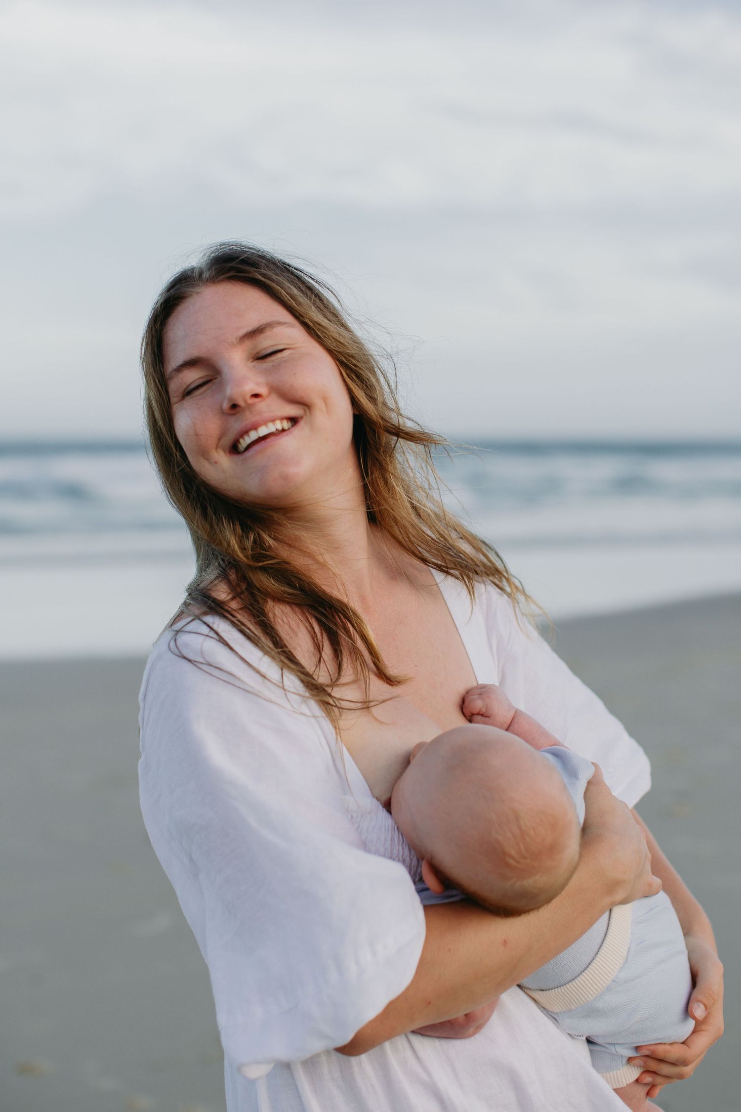 Joyful mother breastfeeding newborn baby on a beach with ocean waves in the background.