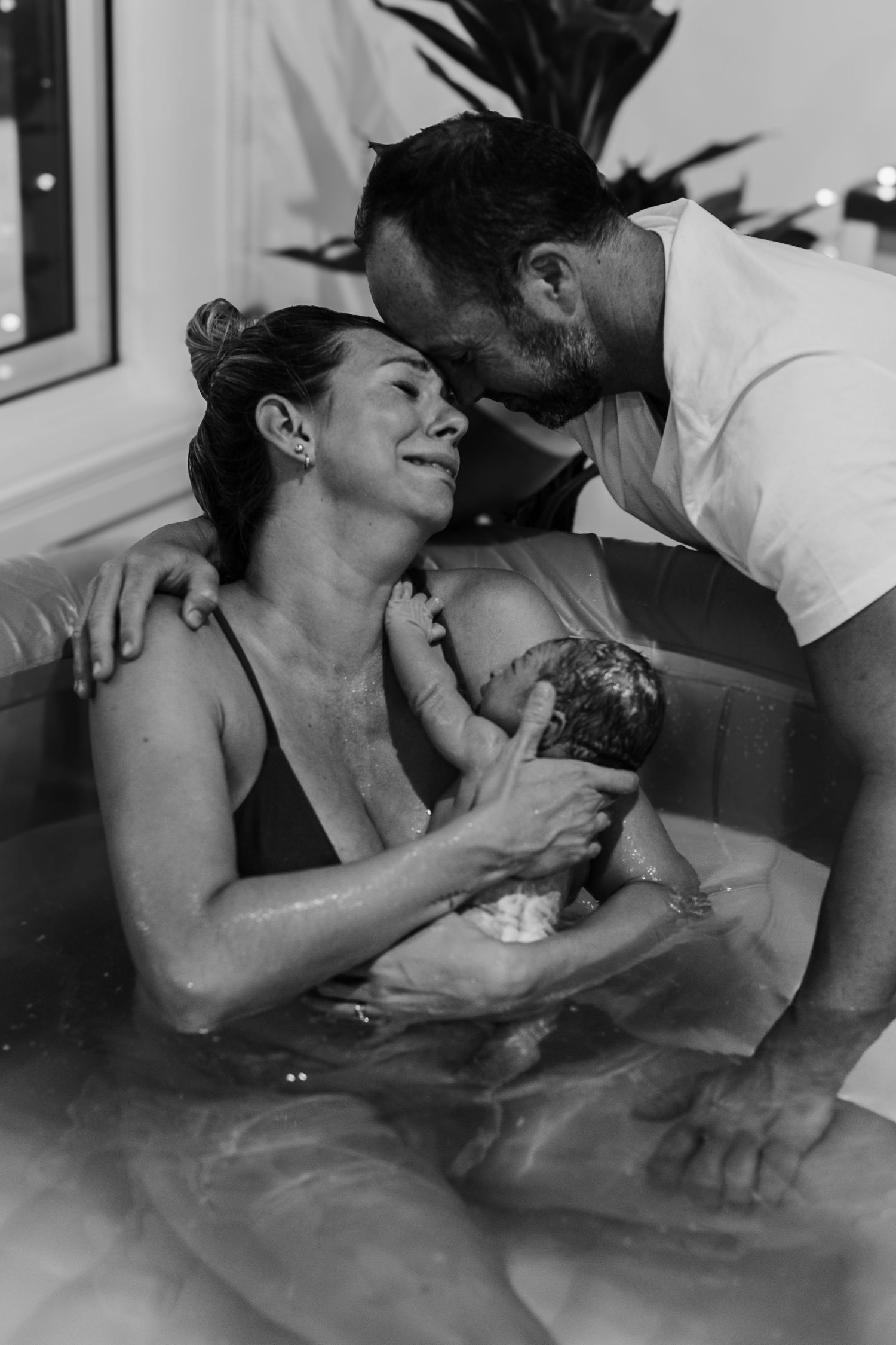 Black and white image of couple sharing a tender moment with their newborn in a water birth pool.