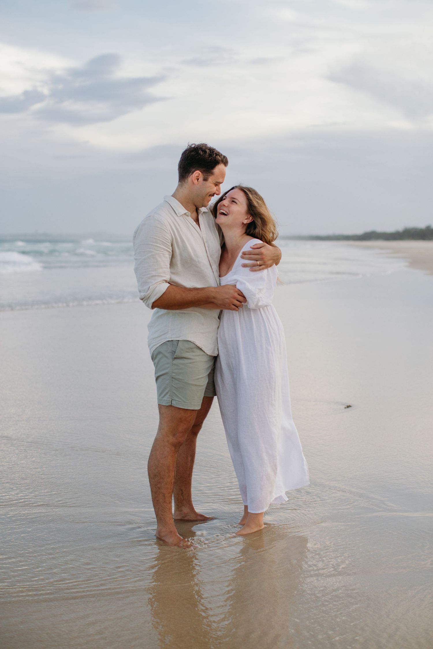 Couple embracing and laughing on a sandy beach at dusk, man in linen shirt and woman in white dress.