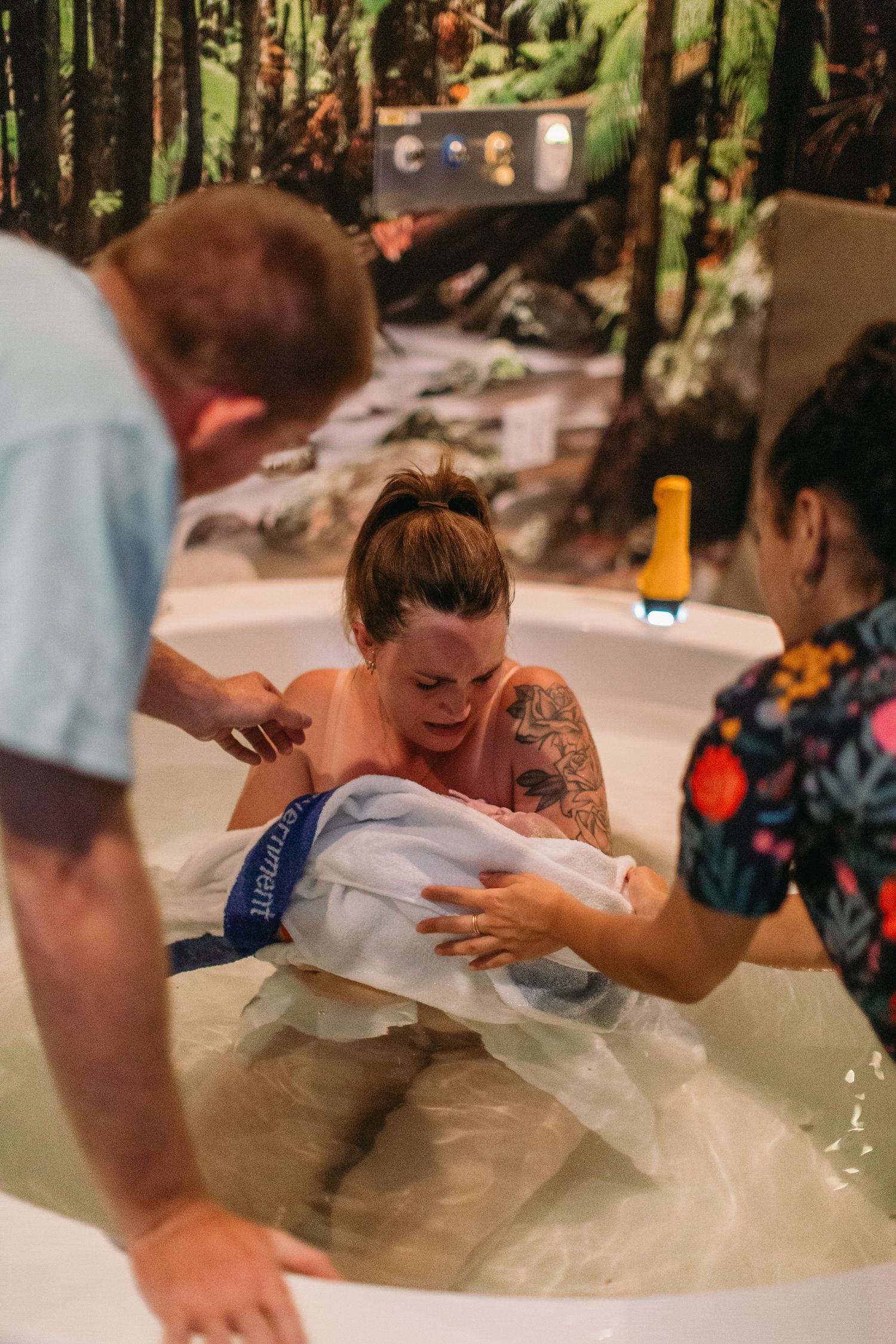 Woman experiencing a water birth in a tub, assisted by two healthcare providers in a nature-themed birthing room.