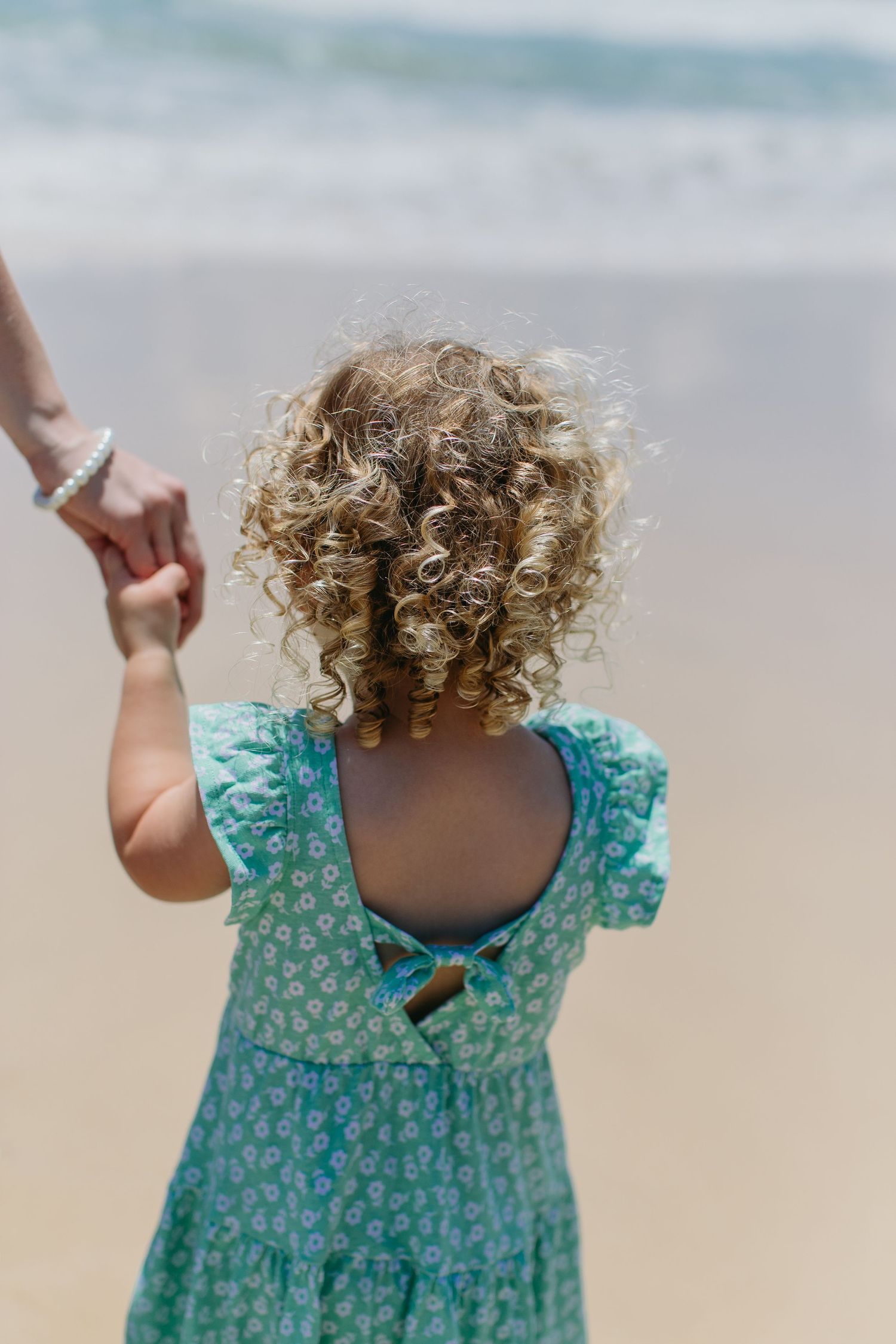 Rear view of curly-haired toddler in teal dress holding mothers hand facing ocean waves.