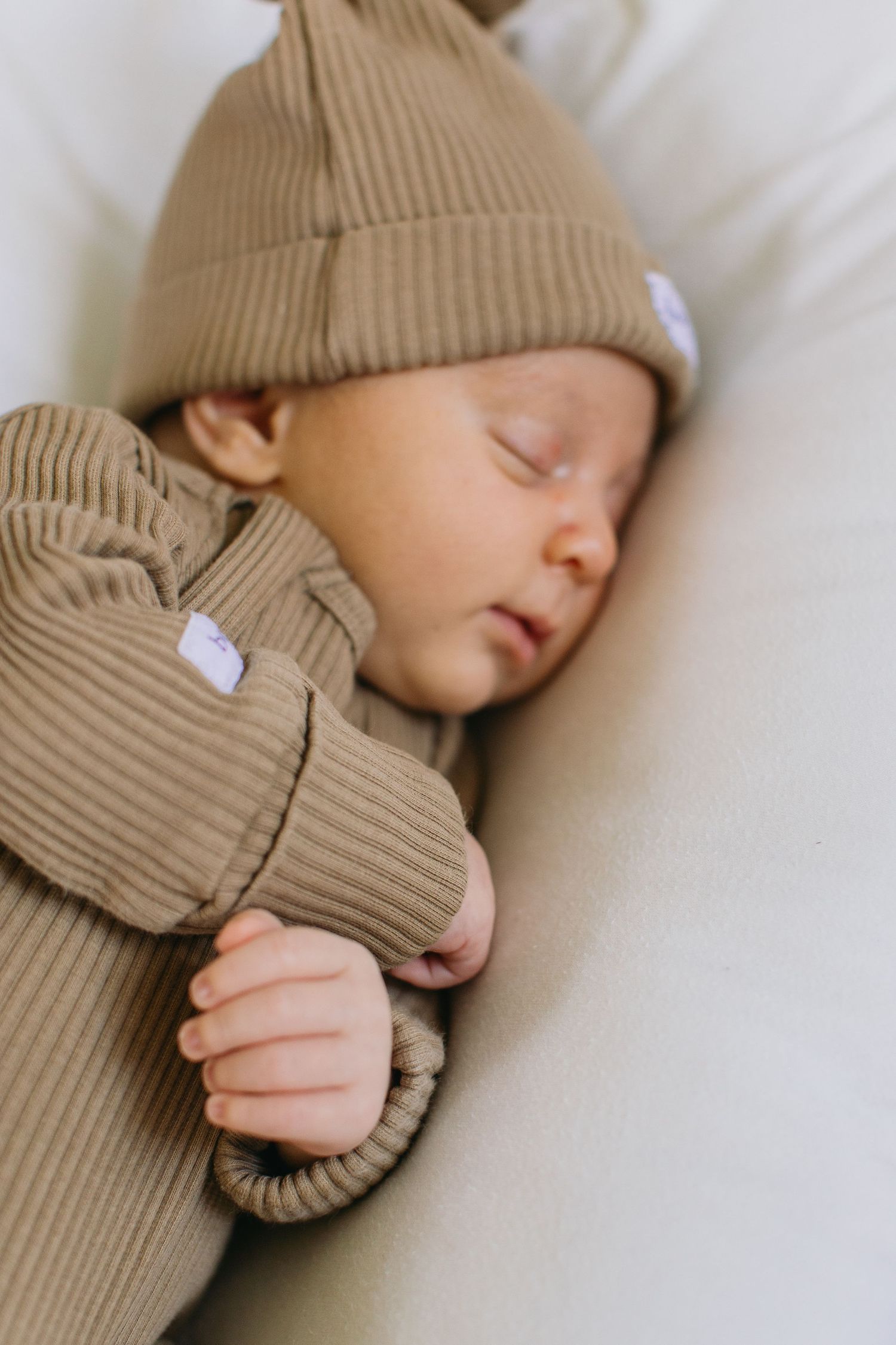 Newborn baby sleeping peacefully in a ribbed tan outfit and matching knot hat on a soft white surface.