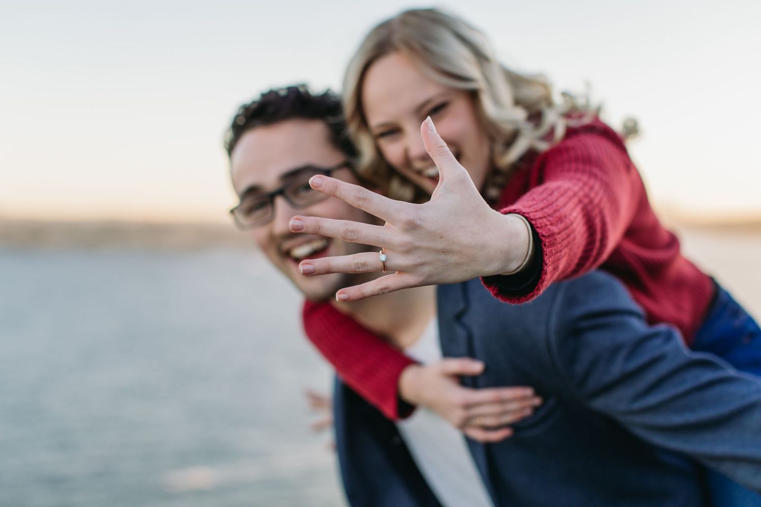 Couple showing off engagement ring outdoors, woman on man's back displaying diamond ring toward camera at sunset.