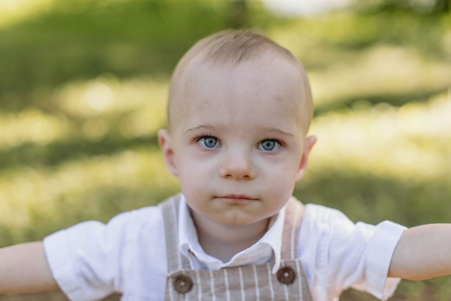 Adorable blue-eyed baby boy in striped overalls outdoors in a sunny green park setting.
