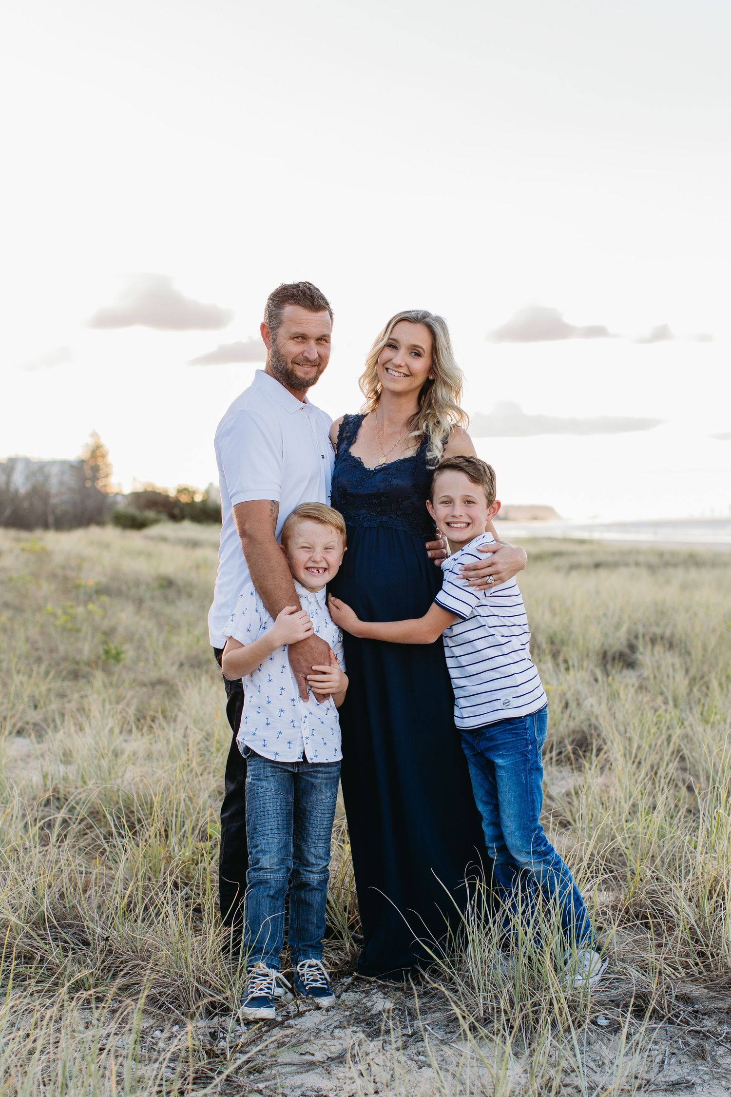 Pregnant woman poses with husband and two young sons in a sunny outdoor field for a family maternity photo.