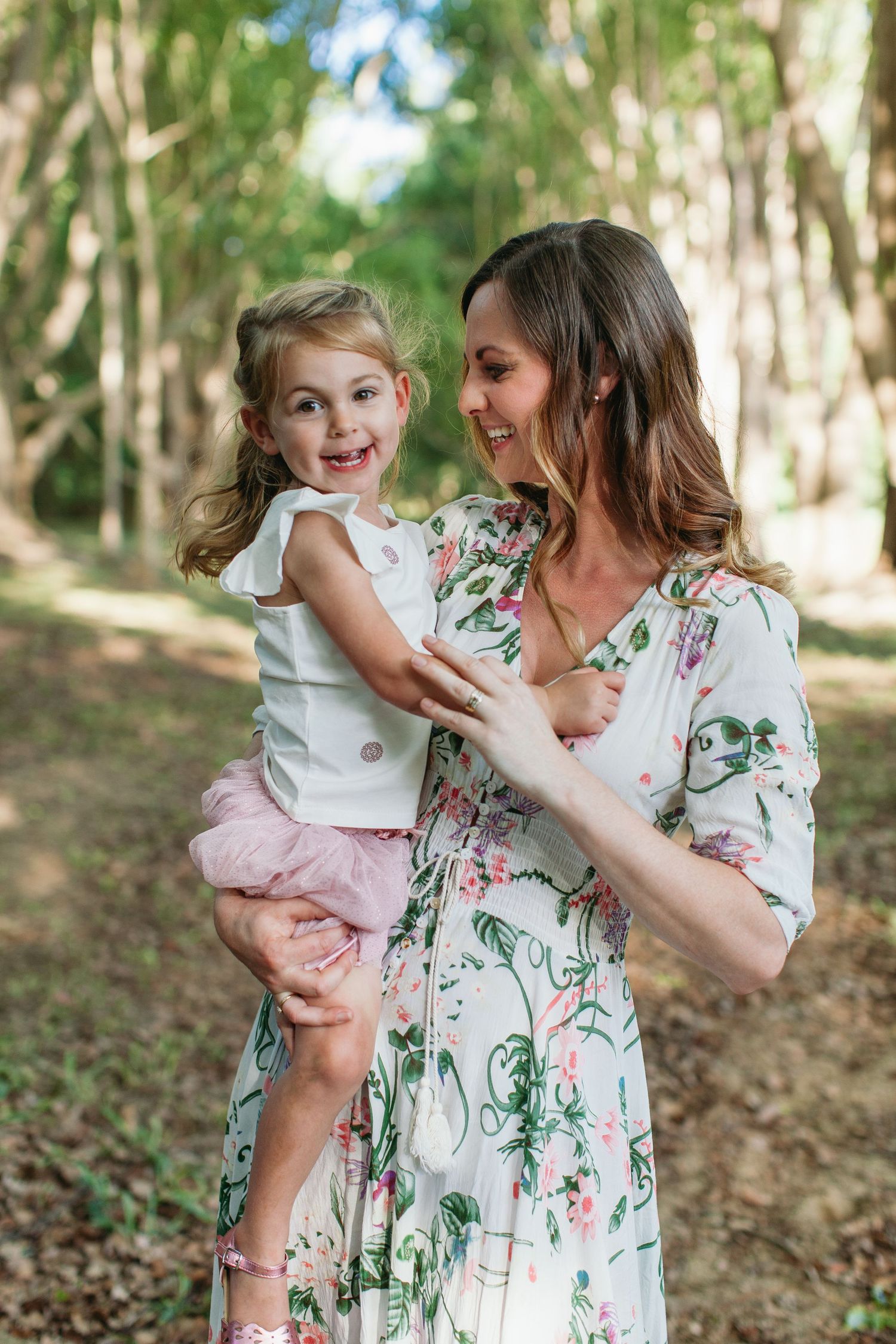A smiling mother in a floral dress holds her happy toddler daughter outdoors in a lush green forest path.