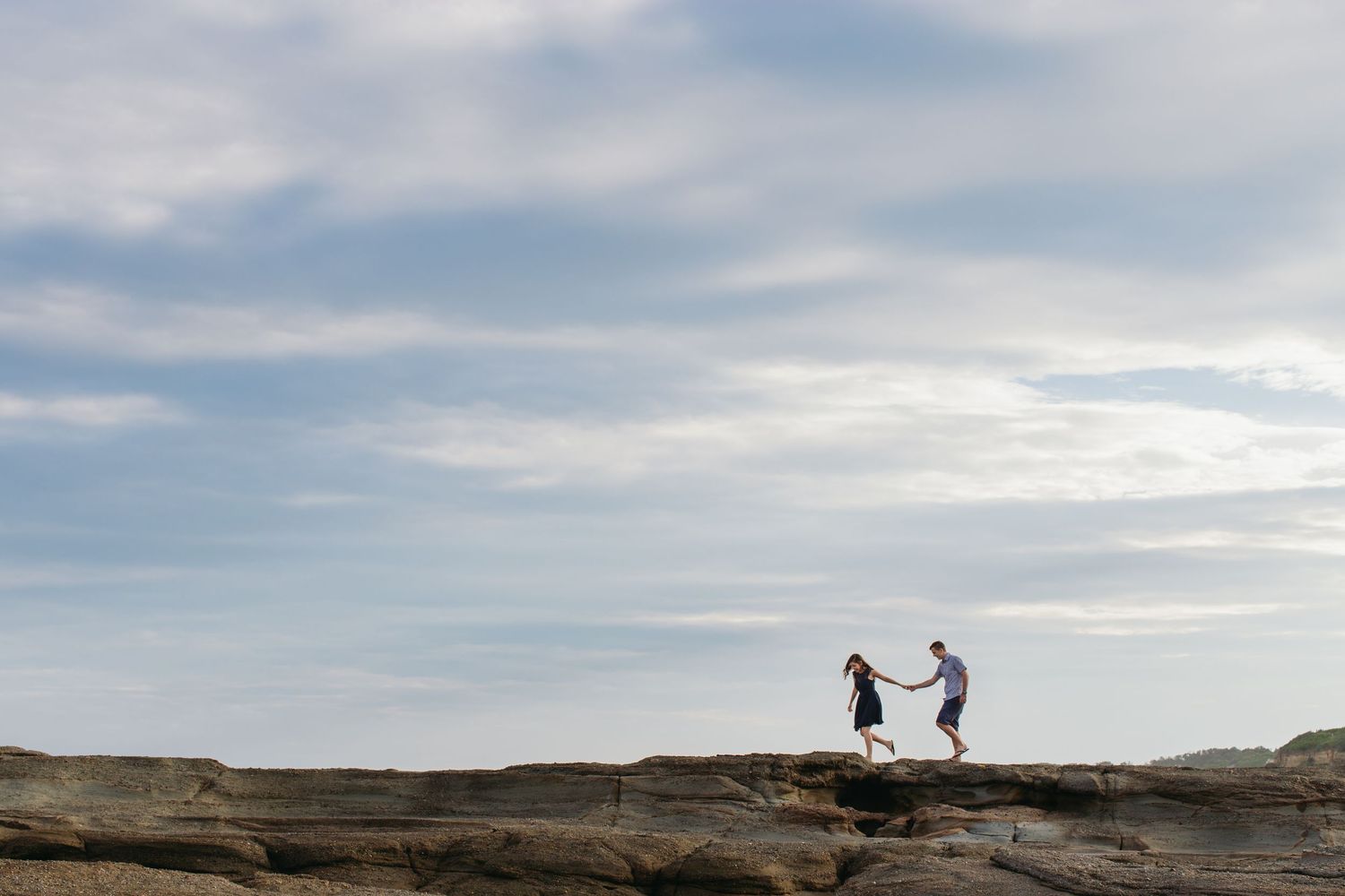 Couple holding hands on rocky coastal cliffs under a vast blue sky at sunset.