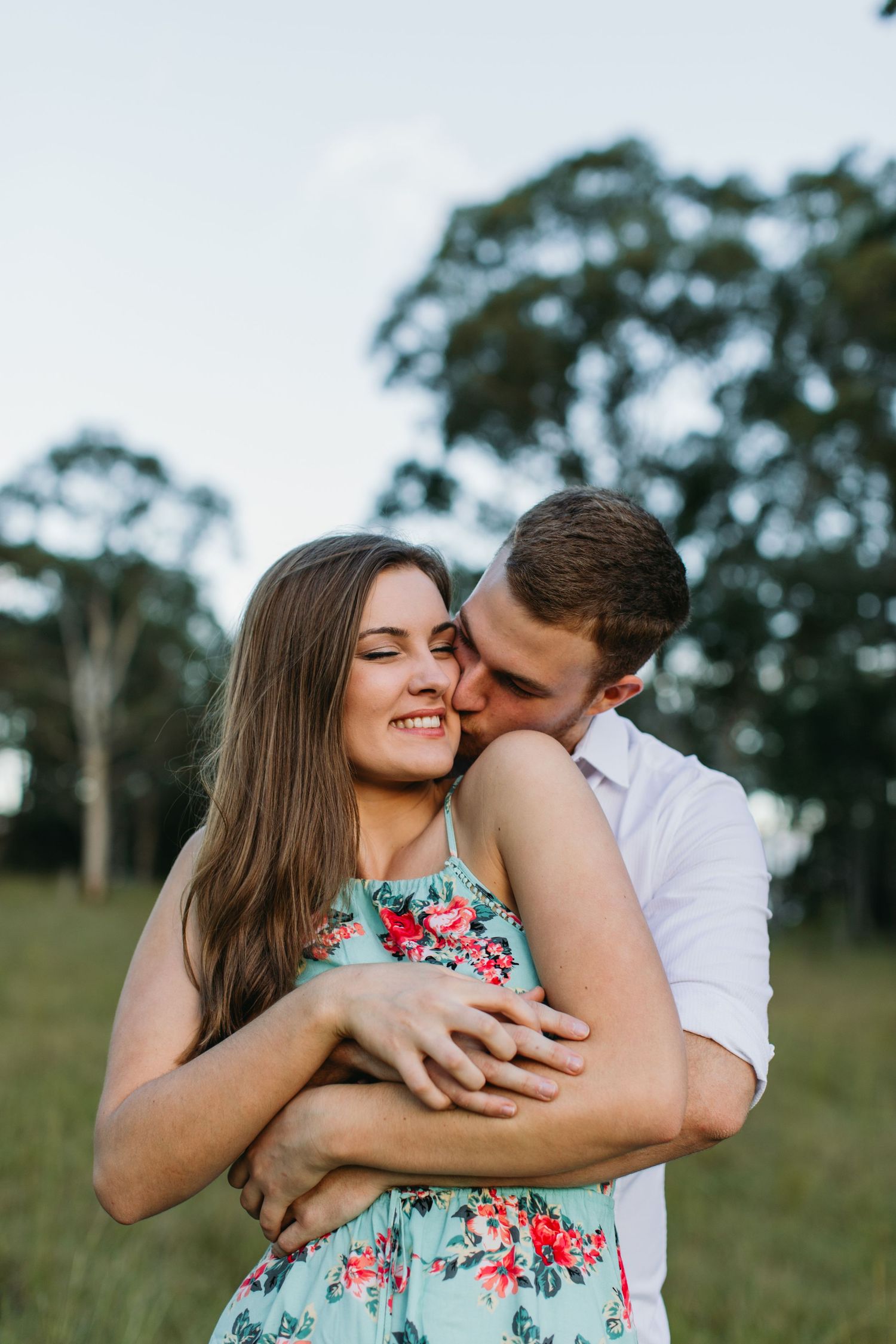 A couple embracing outdoors, woman in floral dress smiling as man kisses her cheek in a green park setting.