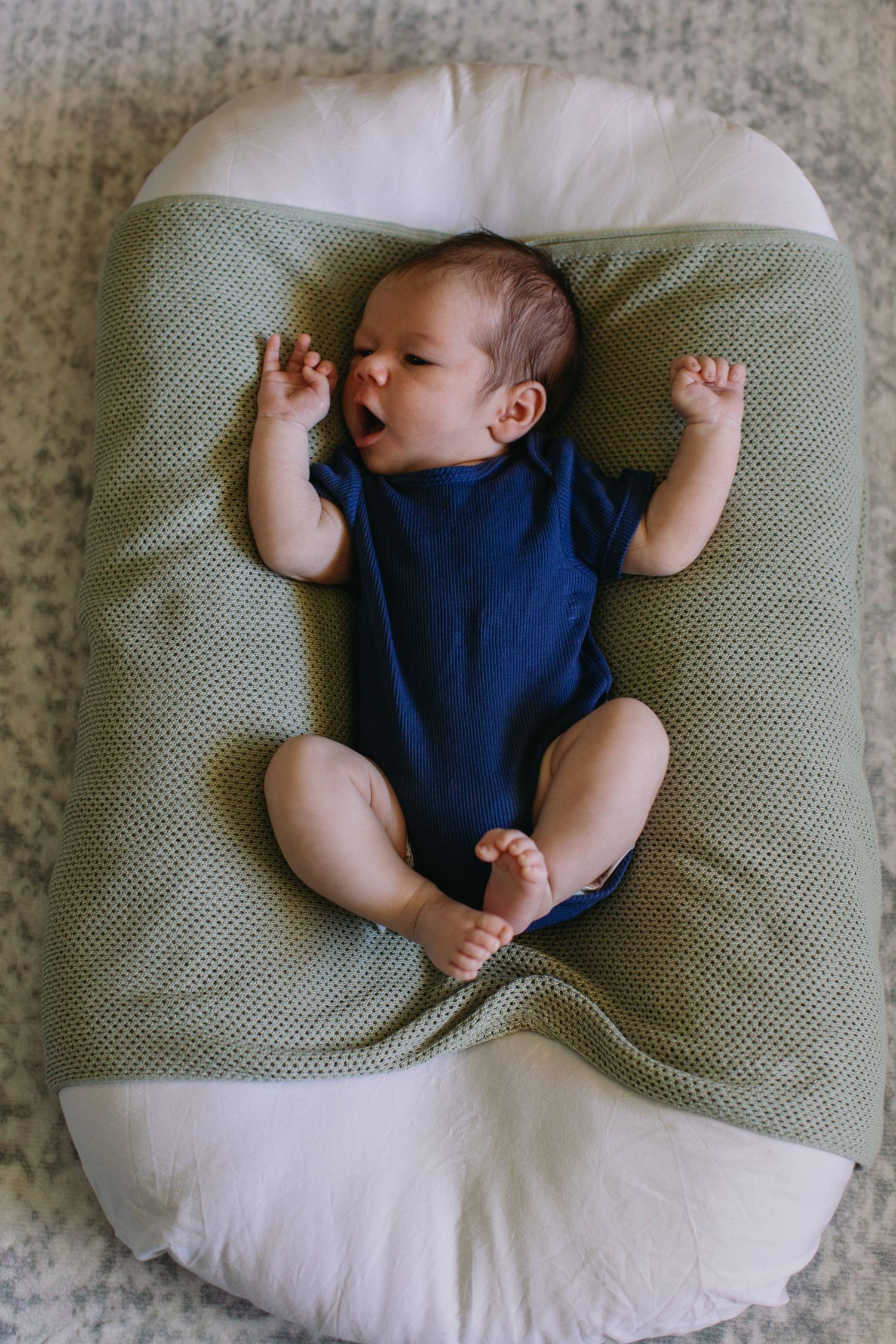 Newborn baby in blue onesie lying on green and white nursing pillow with arms raised and mouth open.
