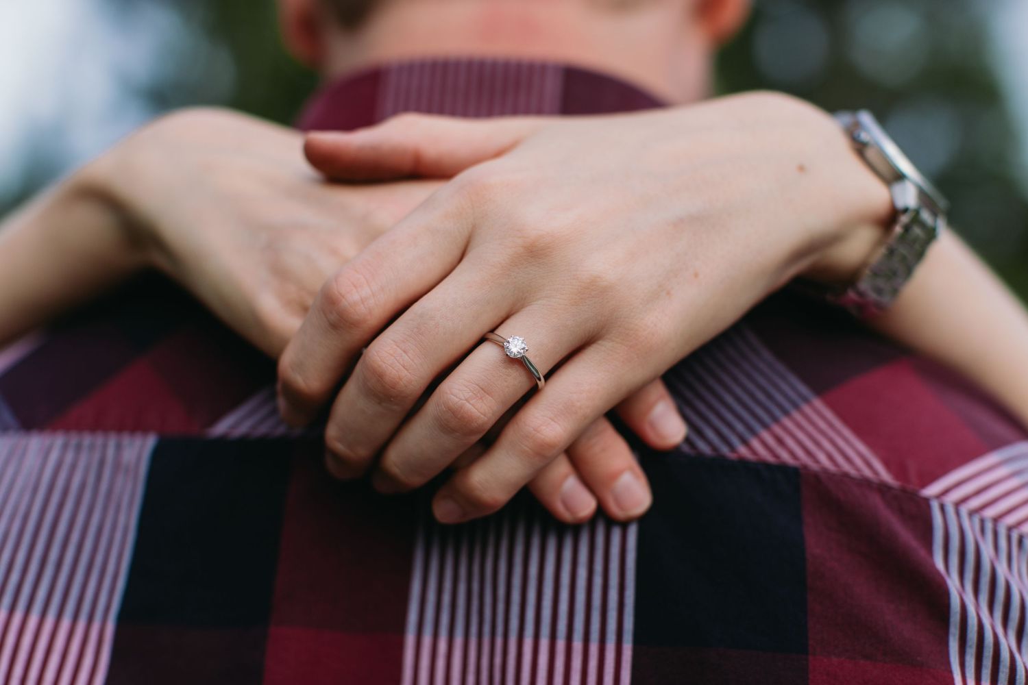 Close-up of a woman's hands with an engagement ring wrapped around a man in a red plaid shirt.