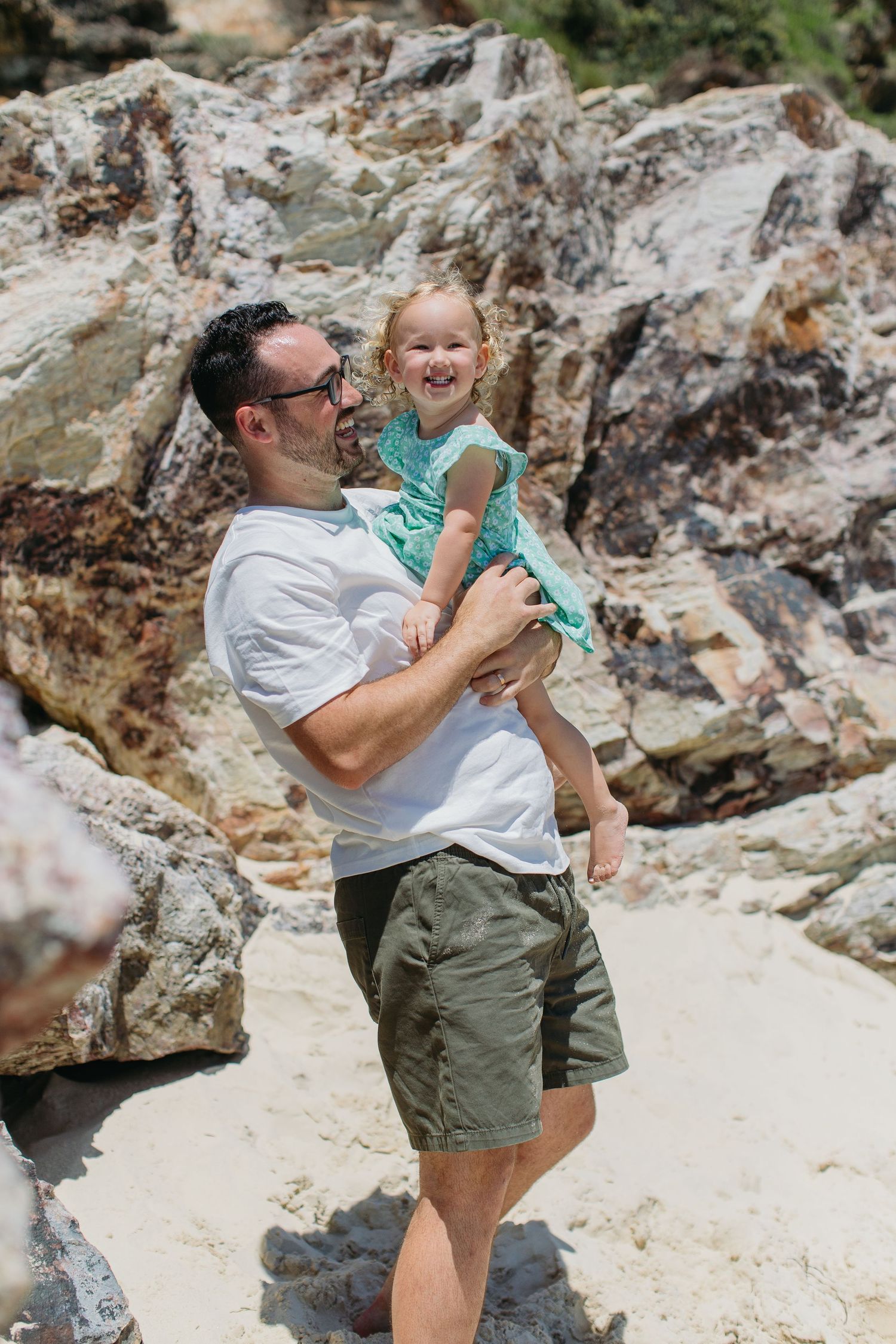 Father holding laughing toddler girl in teal dress at sandy beach with rocky background.