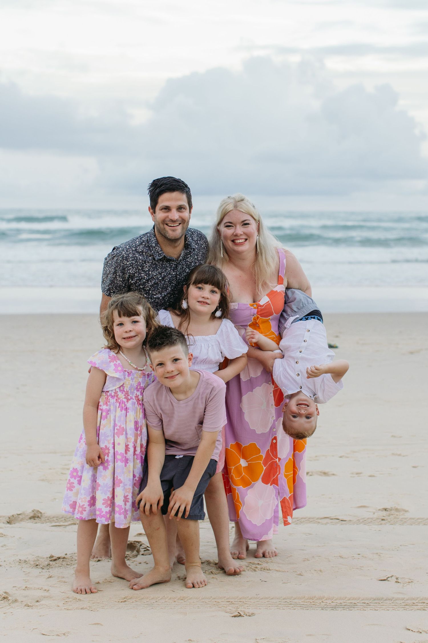 Happy family of six posing together on a sandy beach, parents with four young children smiling at camera.