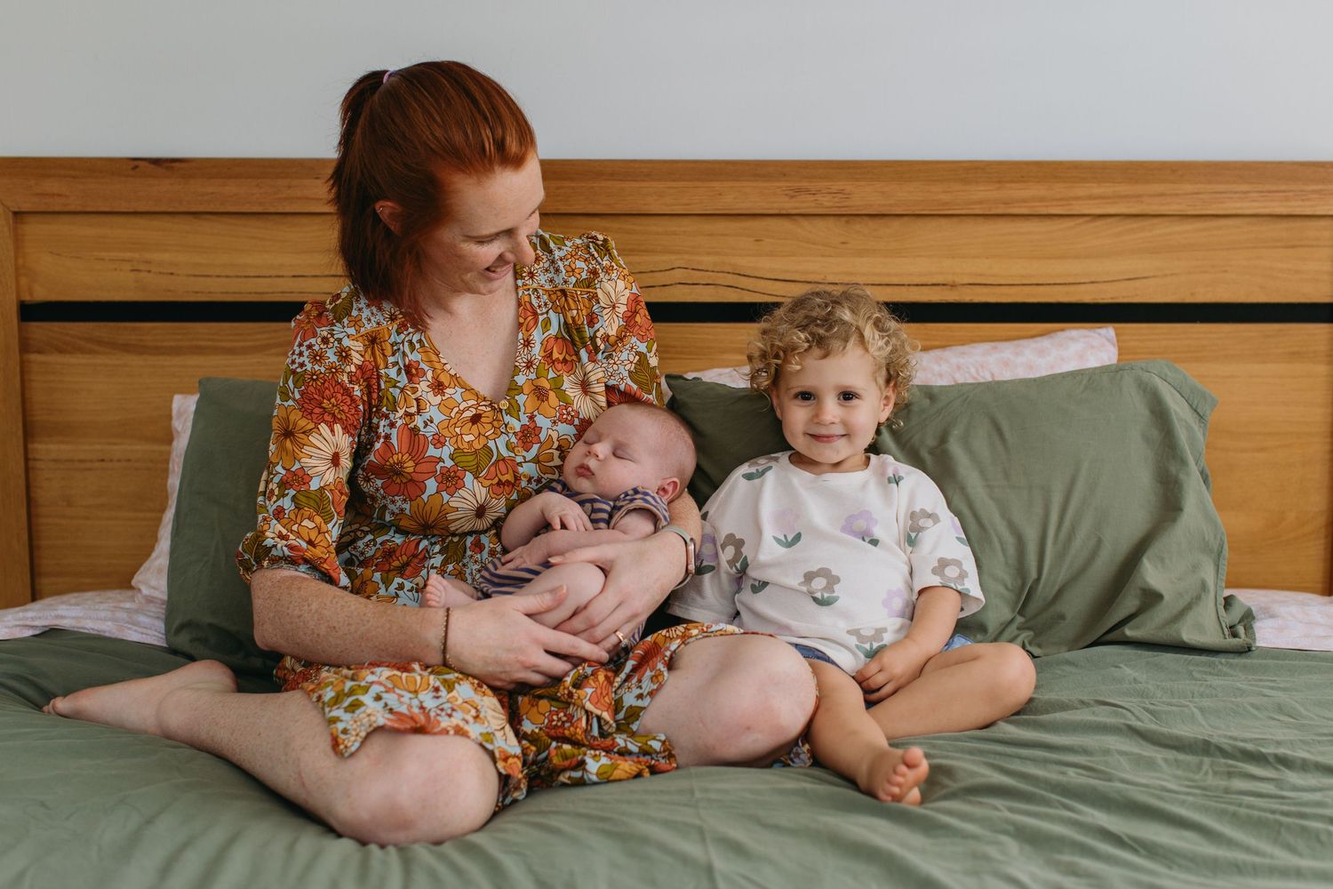 A redheaded mother sits on a green-sheeted bed holding a newborn while her toddler sits beside her smiling.