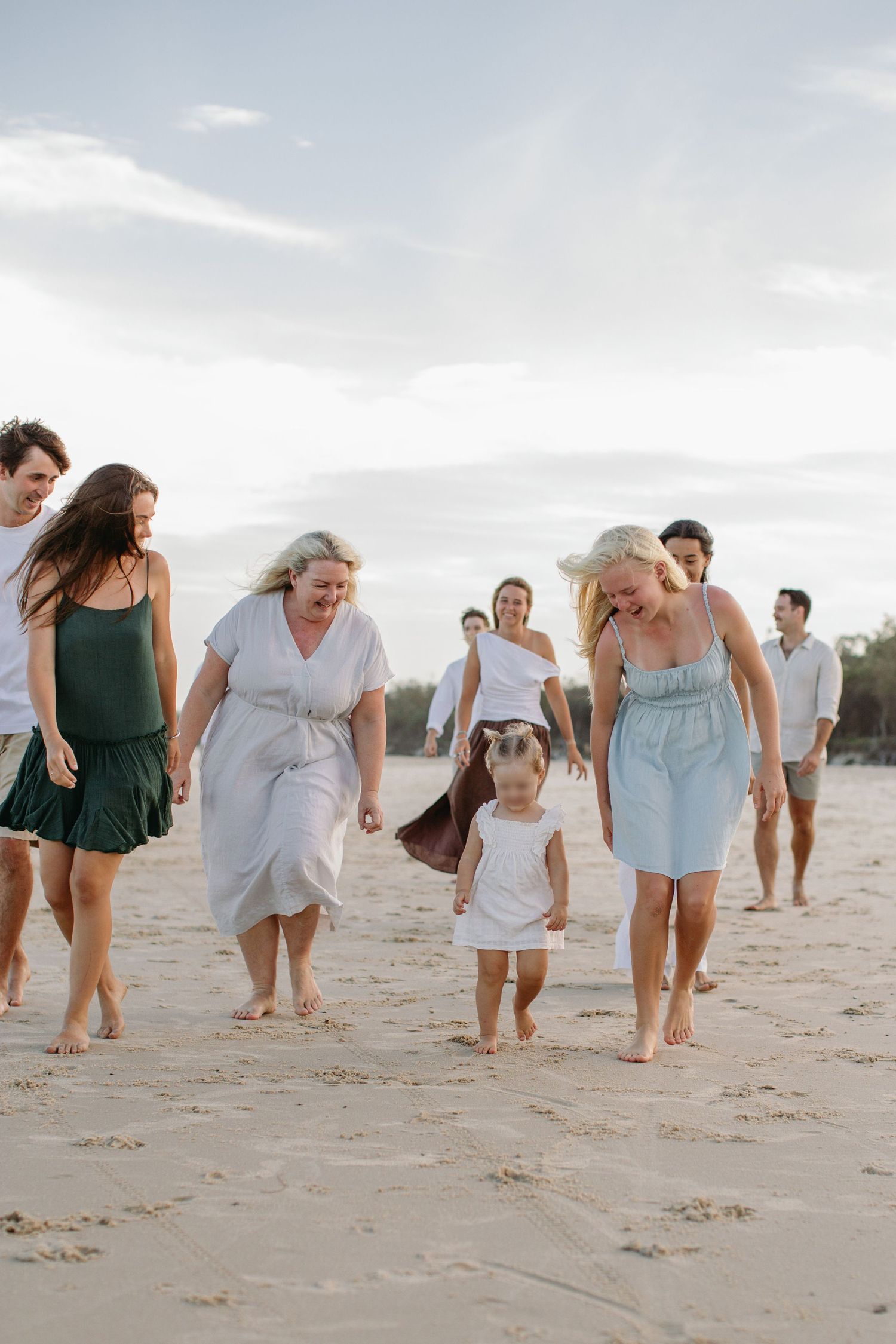 Multi-generational family walking barefoot on sandy beach, smiling and holding hands in casual summer outfits.
