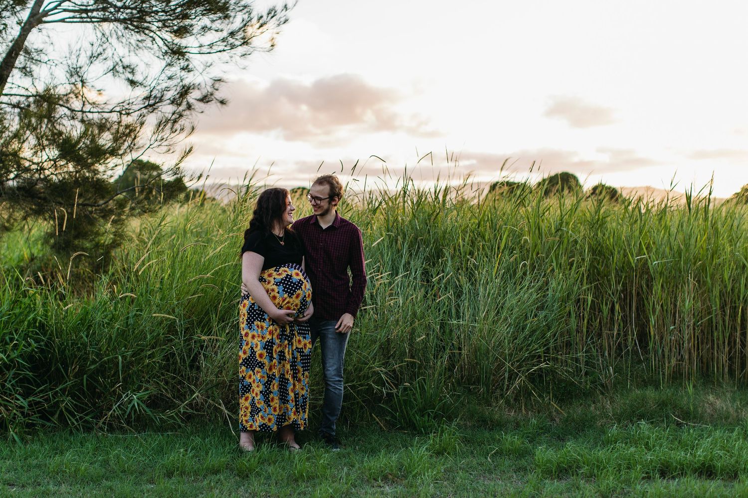 Couple standing in lush green field at sunset, woman in floral dress holding flowers, man in burgundy shirt smiling.