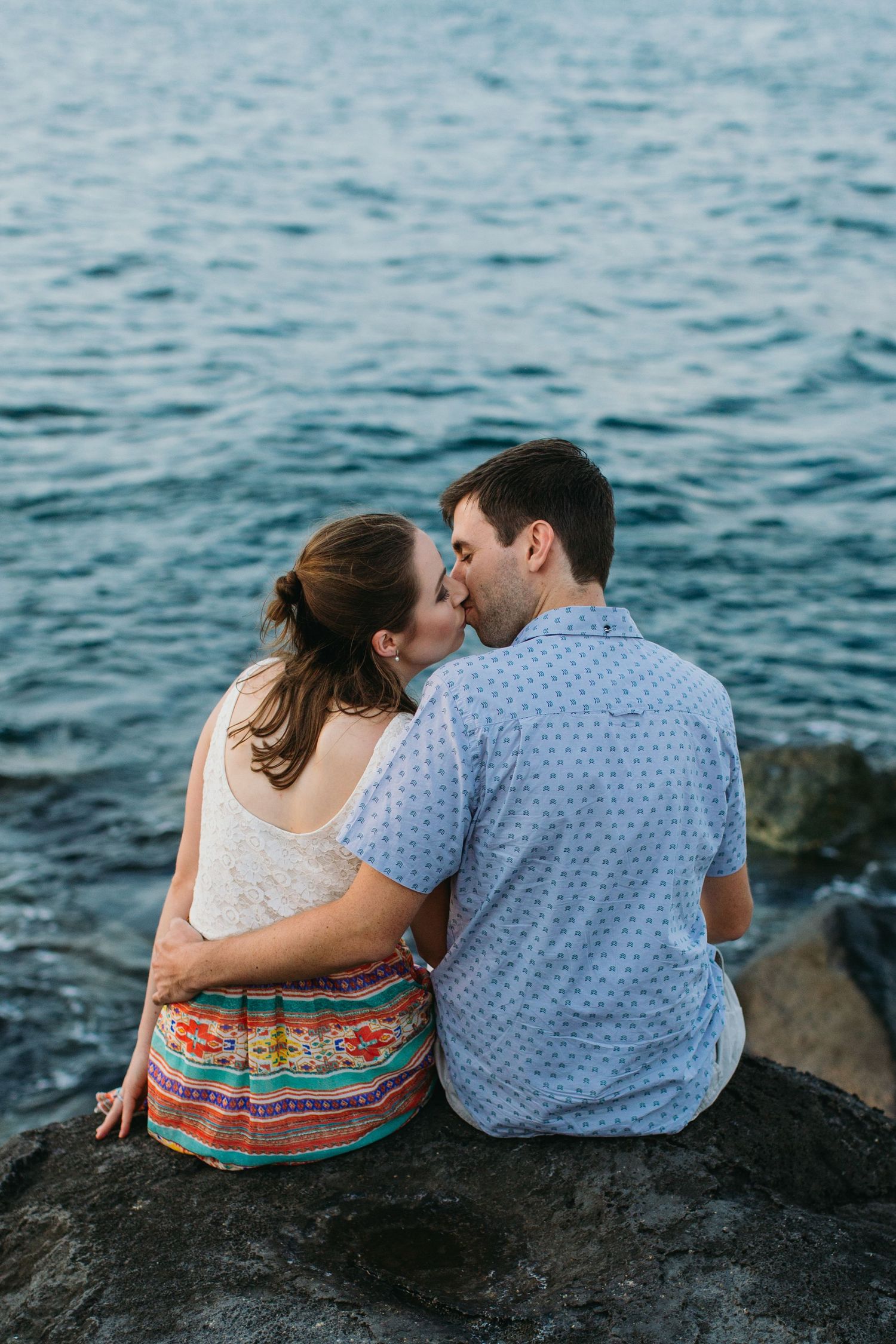 Couple sharing a romantic kiss while sitting on rocks by the water, woman in colorful skirt.