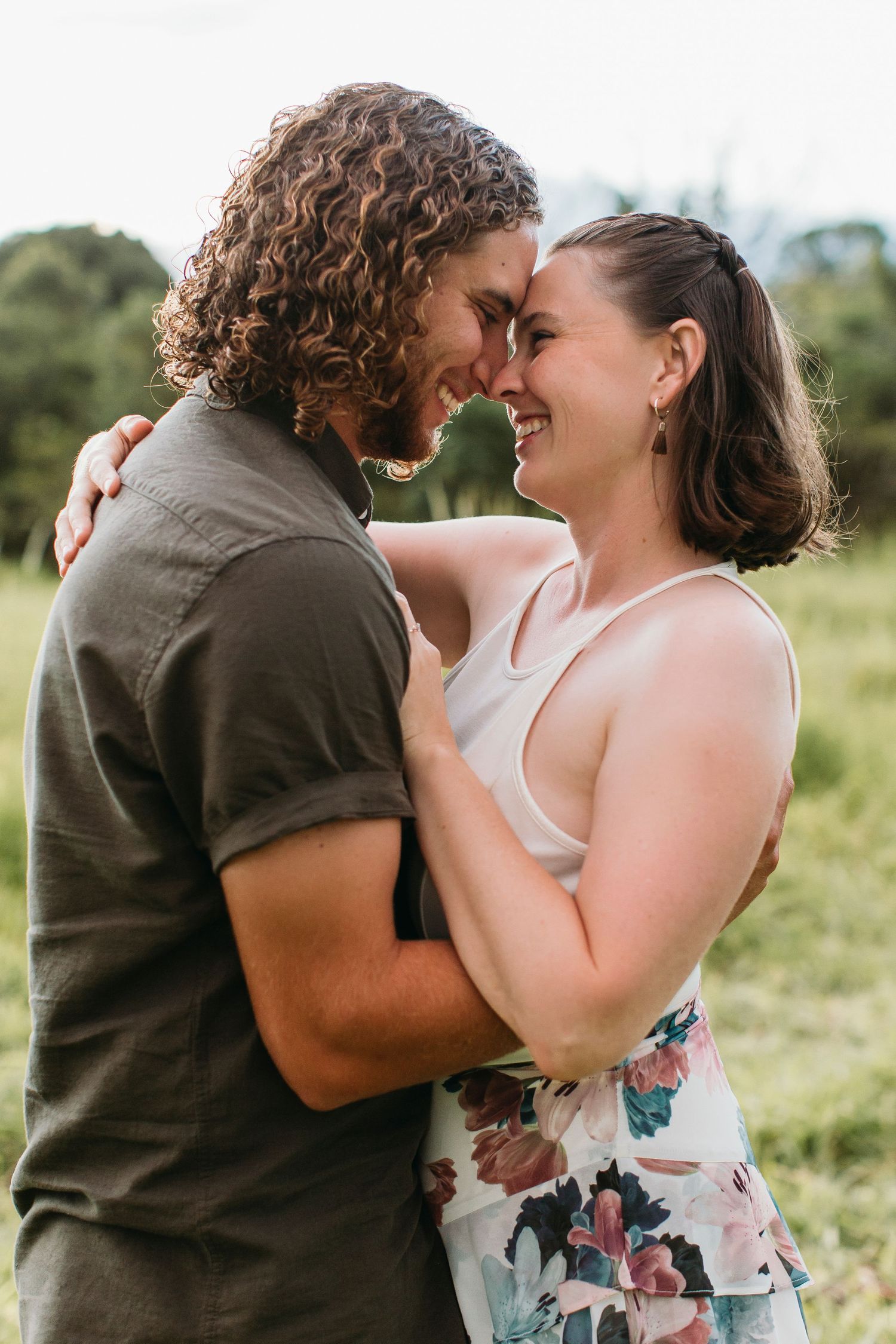 A couple embraces outdoors, touching foreheads and smiling in a romantic moment surrounded by greenery.