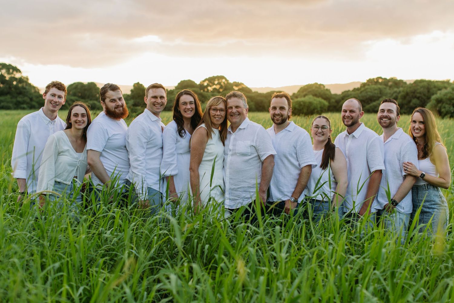 Group of smiling adults in white shirts posing in a lush green field at golden hour sunset.