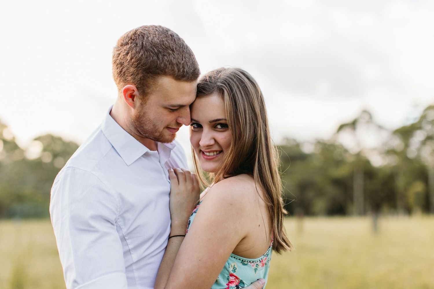 A couple embraces in a sunny field, the woman smiling at the camera in a floral dress.