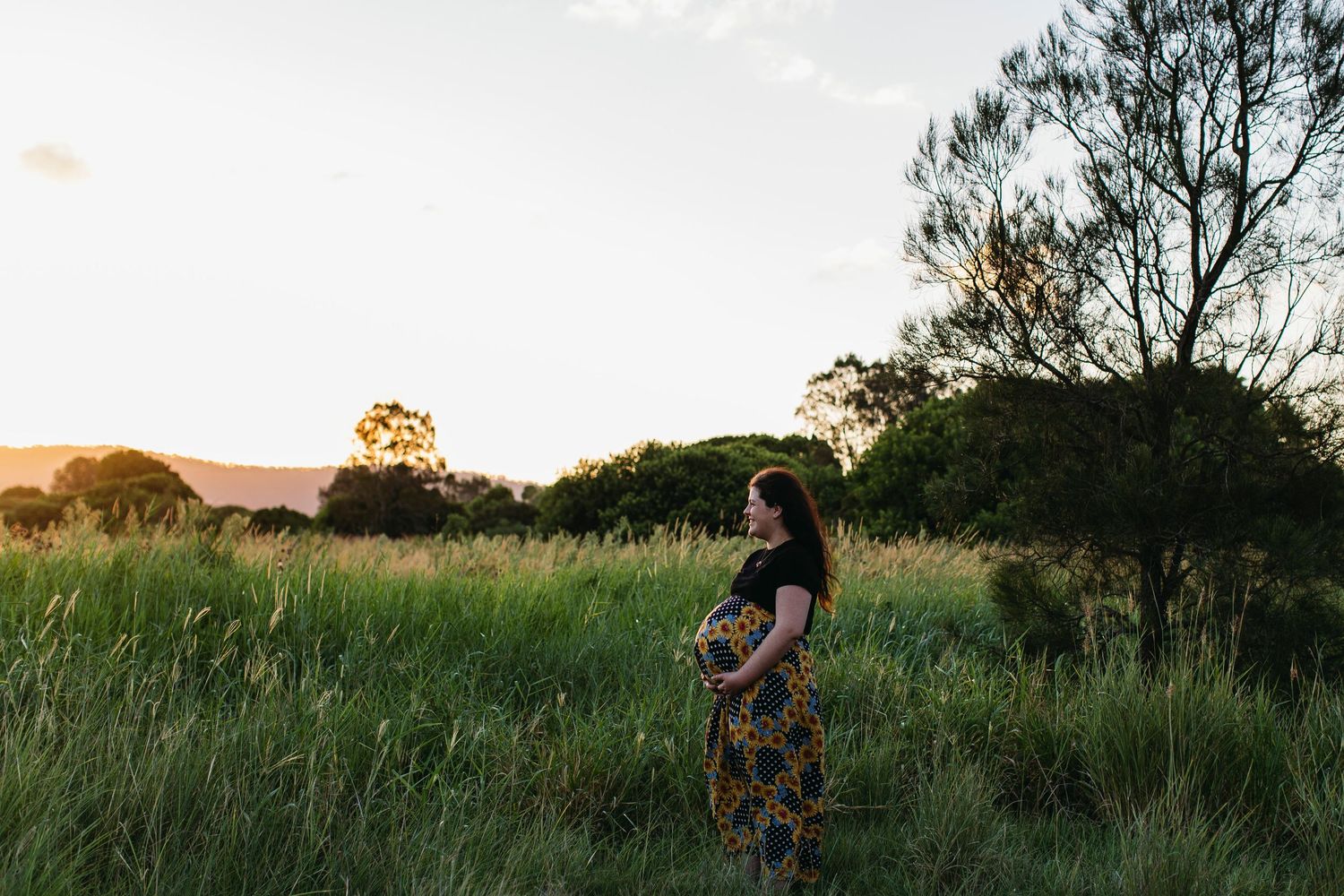 Pregnant woman in floral skirt cradling her baby bump in a lush green field at golden sunset.