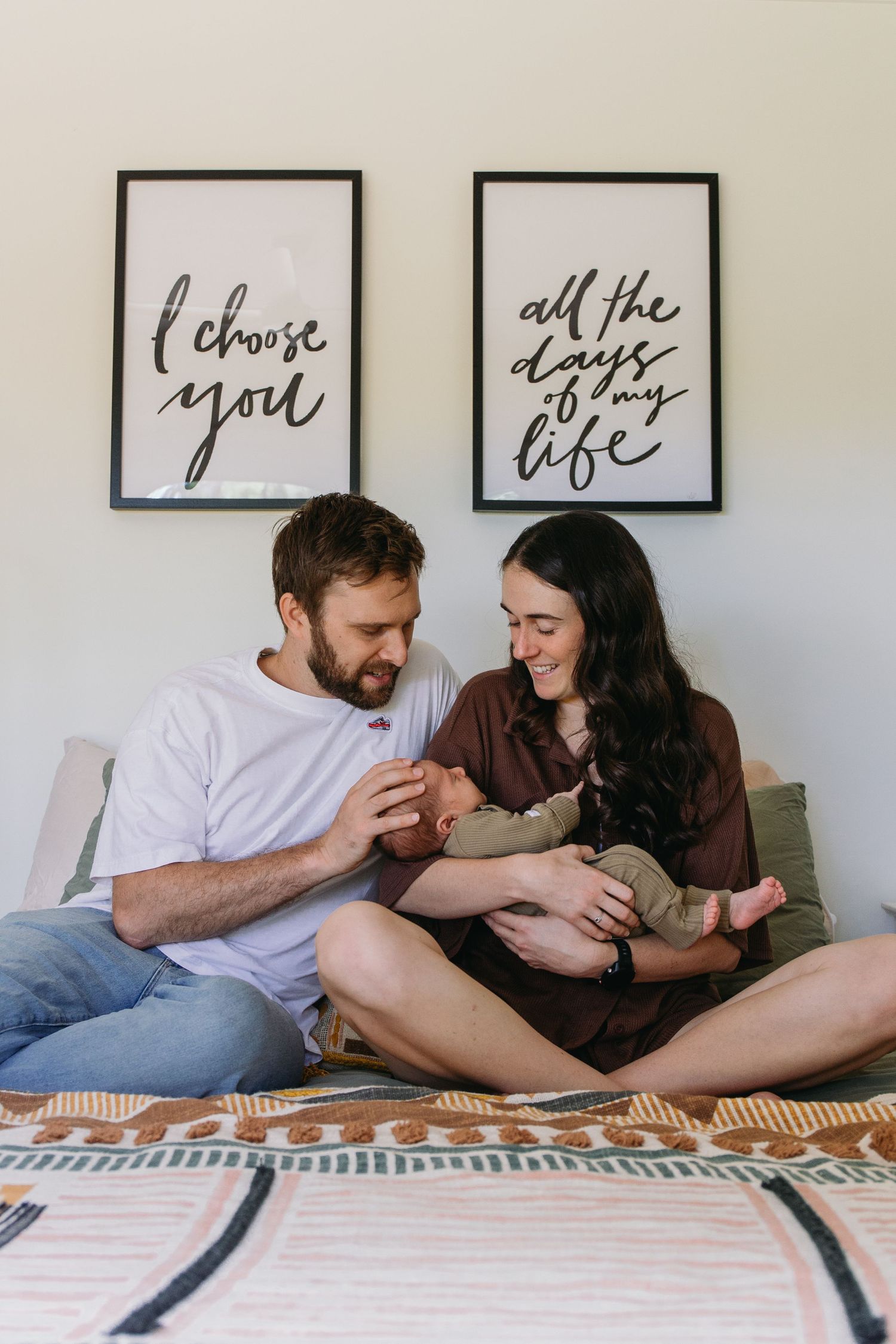 New parents sitting on bed smiling at newborn baby, with romantic wall art reading I choose you all the days of my life.