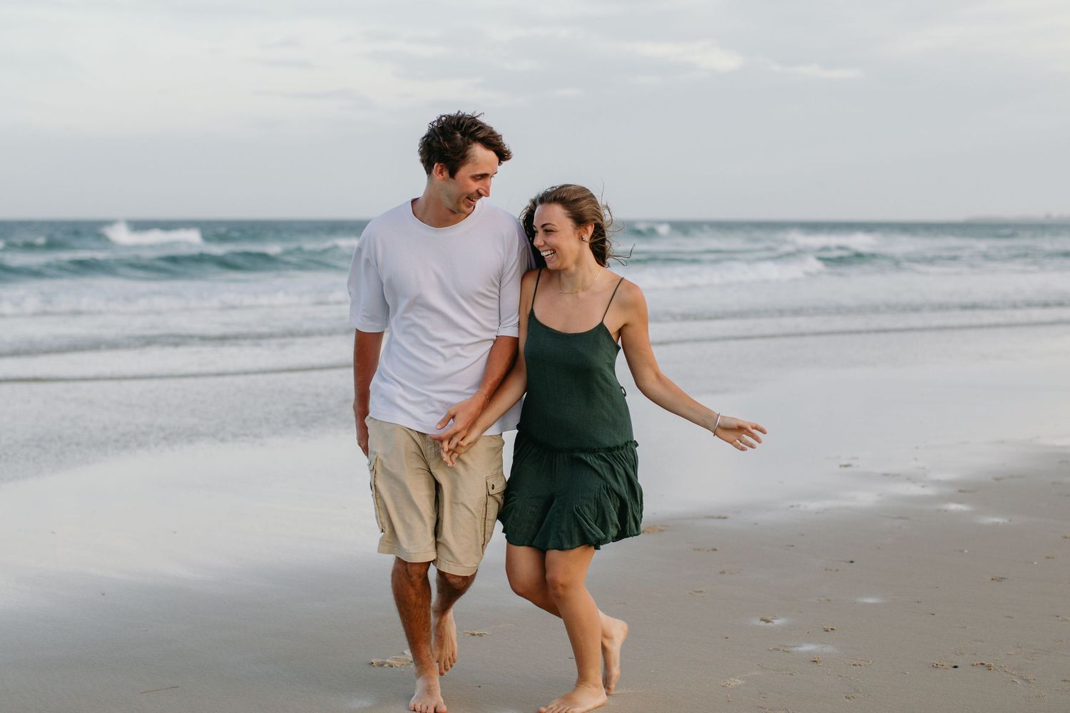 Couple walking hand-in-hand on a sandy beach, smiling at each other with ocean waves in the background.